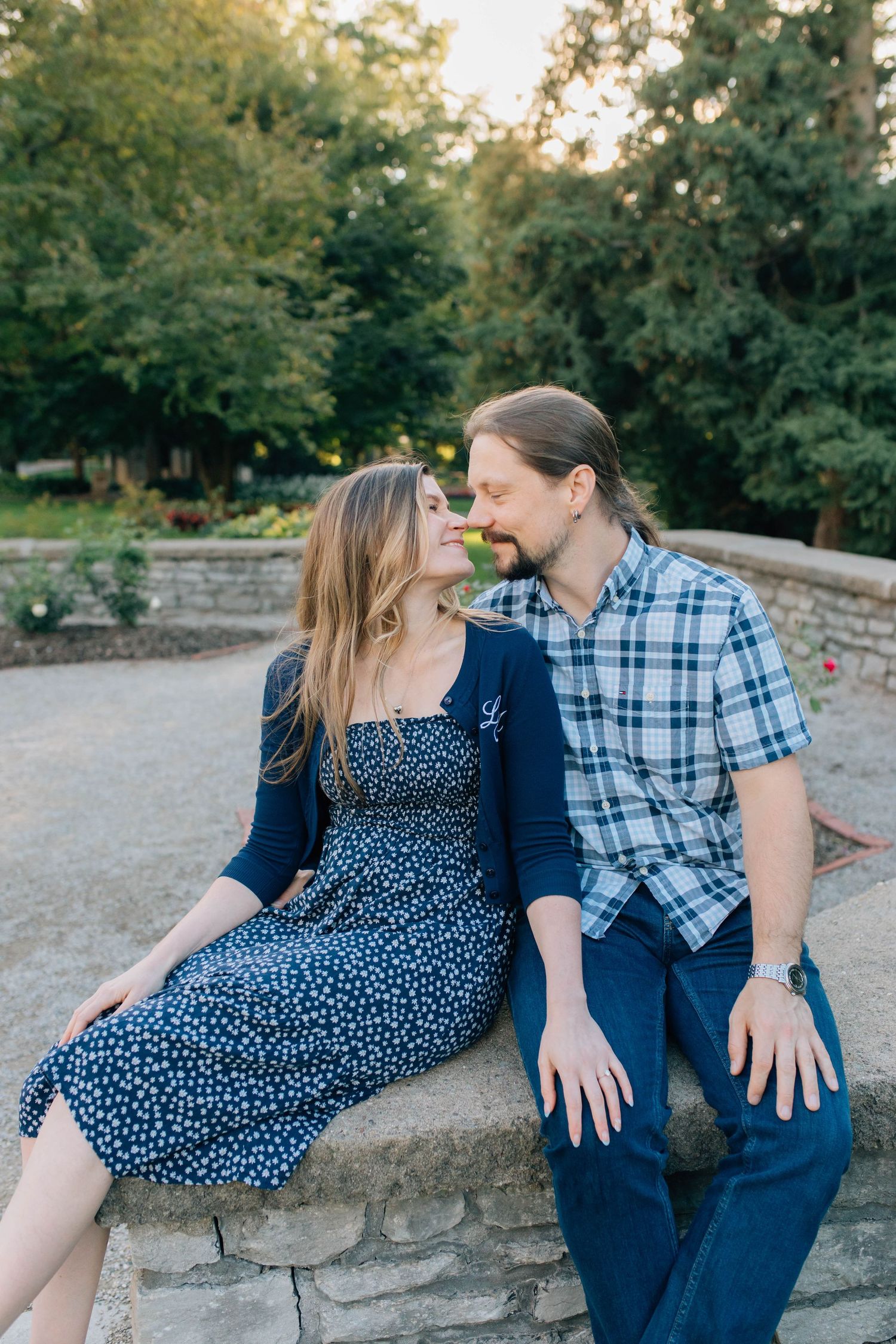 Couple sharing a quiet romantic moment seated on stone wall in lush garden, woman in navy floral dress.