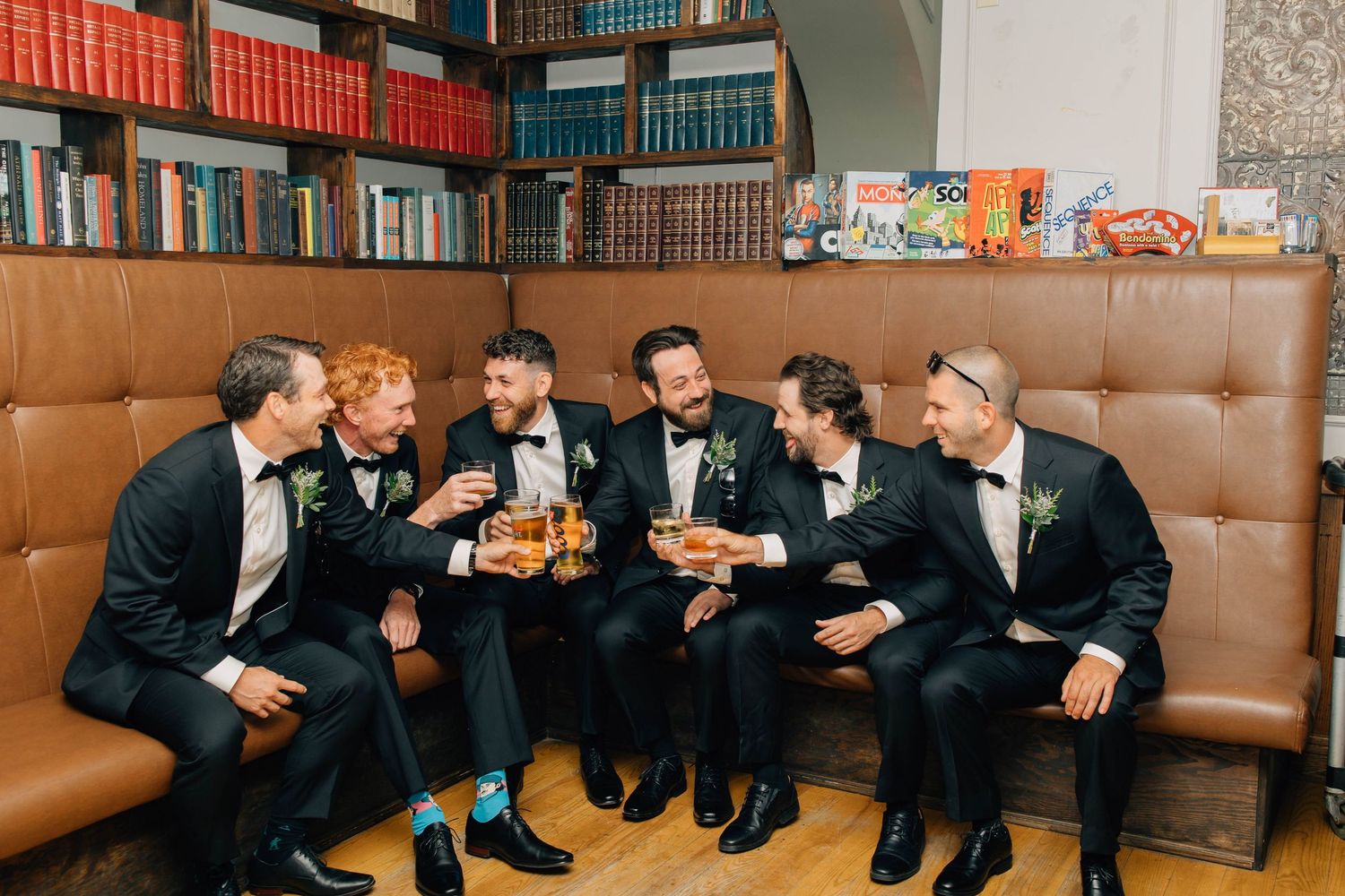 Groomsmen in black tuxedos celebrate with drinks and snacks on a leather sofa in a library setting.