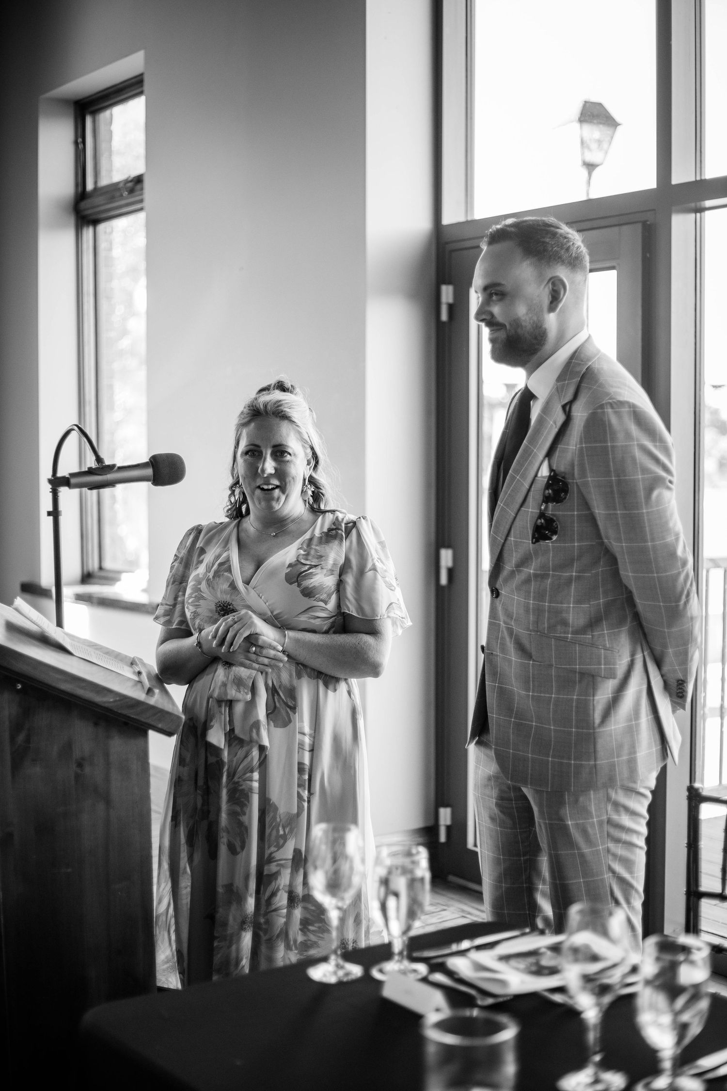 Woman giving speech at podium beside groom in plaid suit at wedding reception in black and white photo.