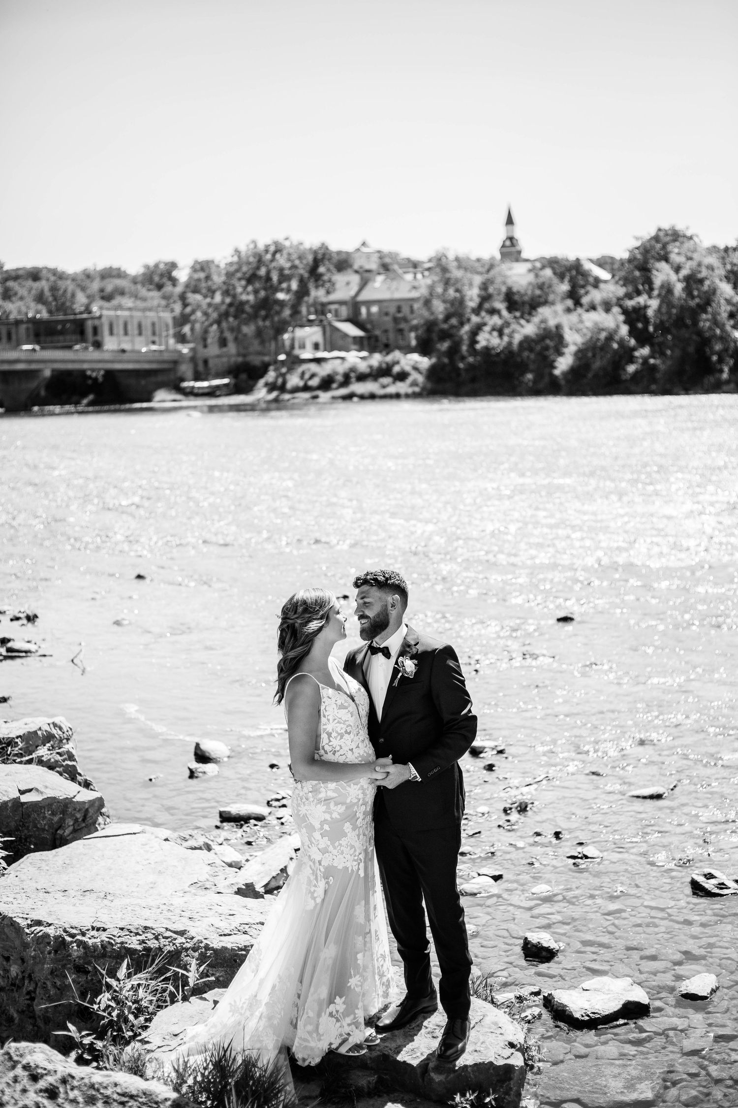 Black and white photo of bride and groom embracing by a sparkling river with a church steeple in the background.