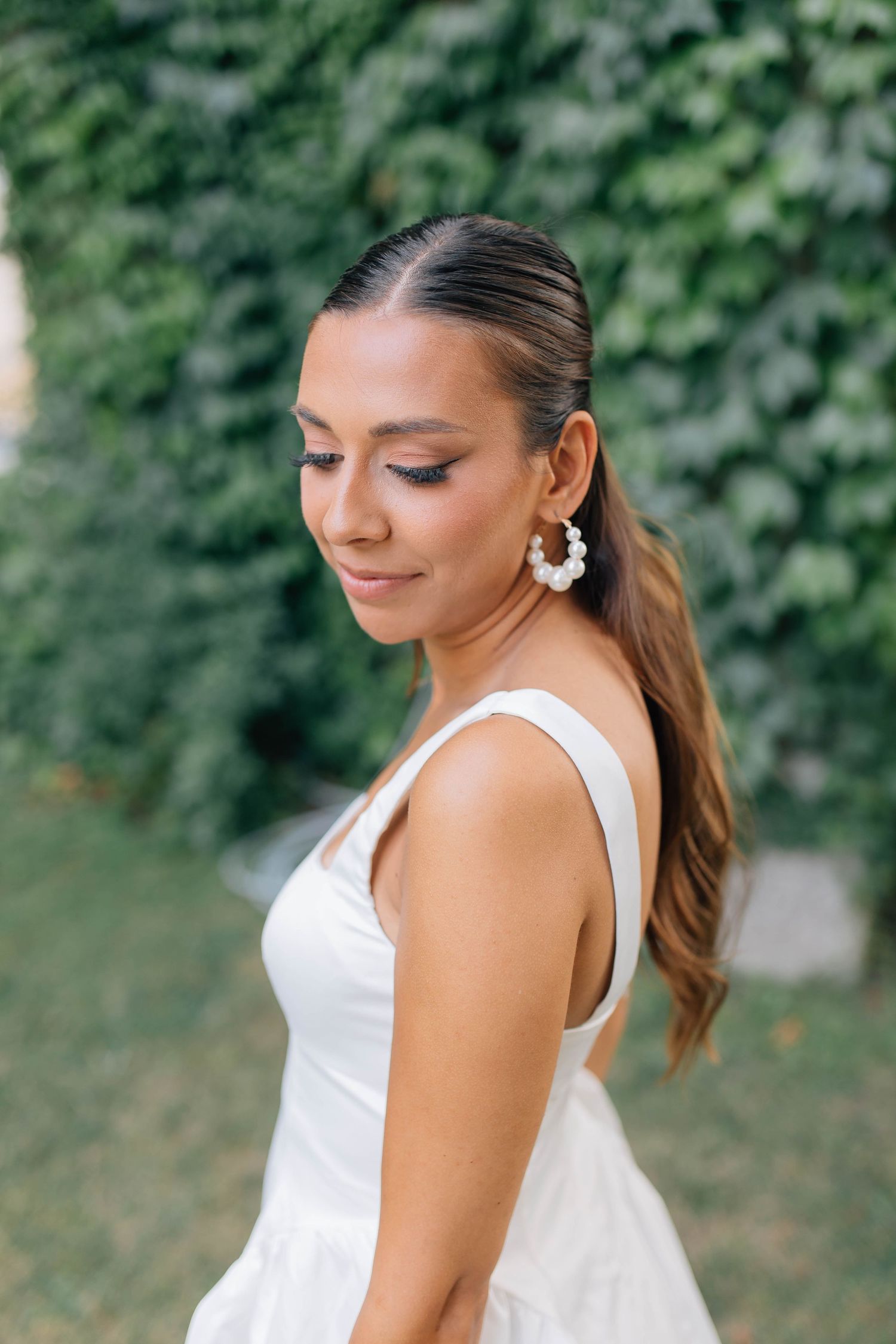Elegant bride in white dress with floral earrings posing in front of a lush green ivy-covered wall.