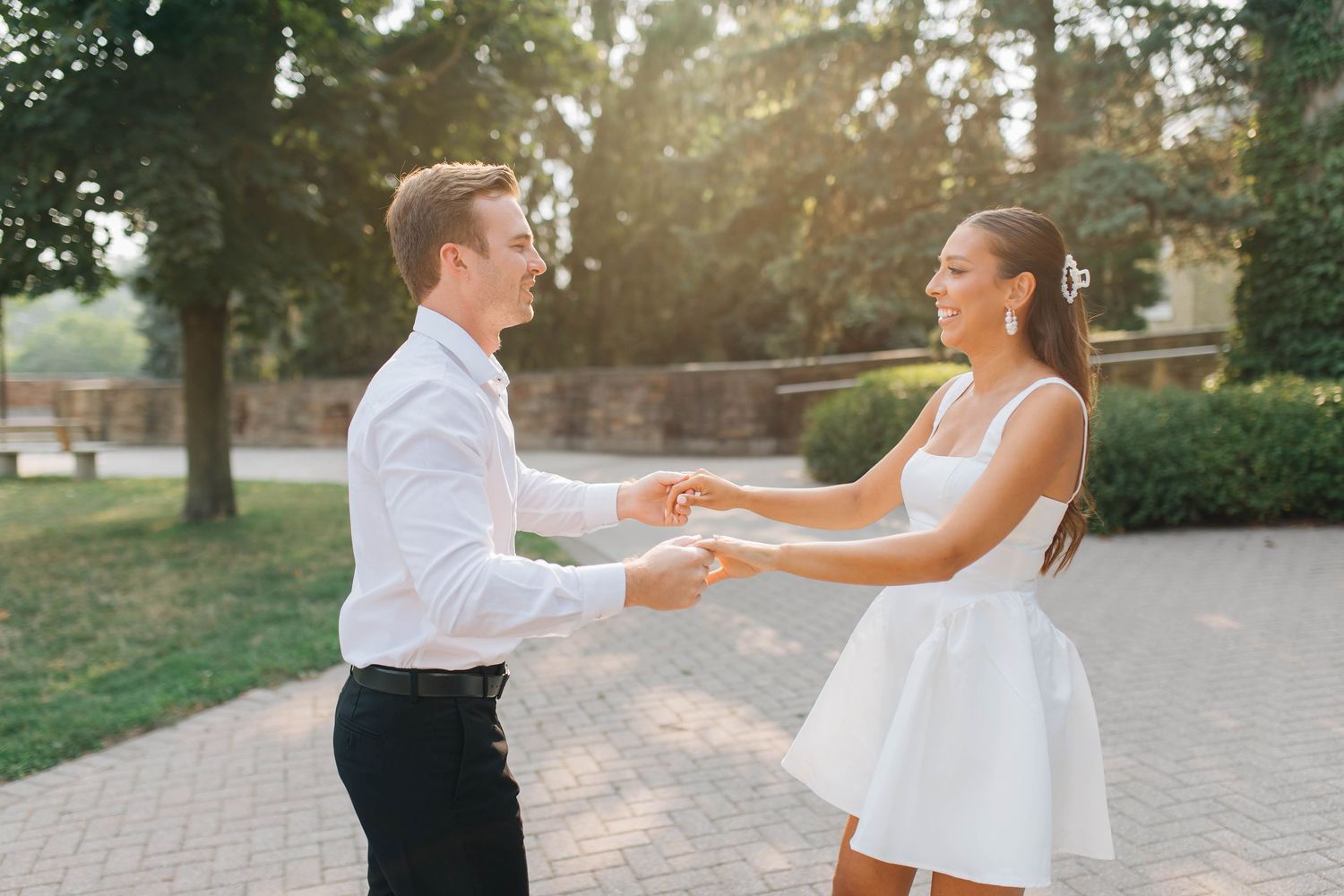 Bride and groom holding hands facing each other on a sunlit brick path surrounded by green trees.