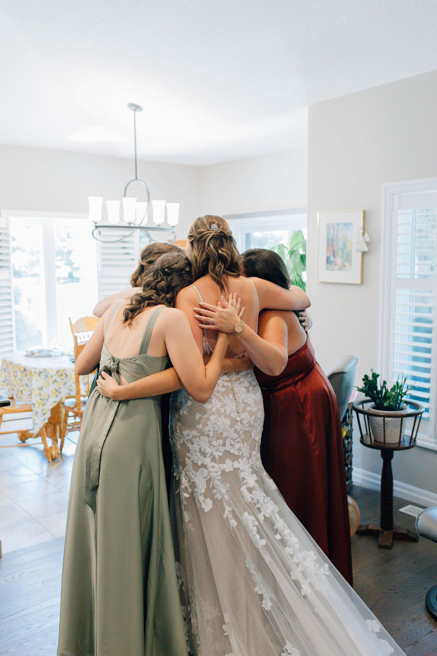 Bride in lace wedding gown shares an emotional group hug with bridesmaids in sage and burgundy dresses.