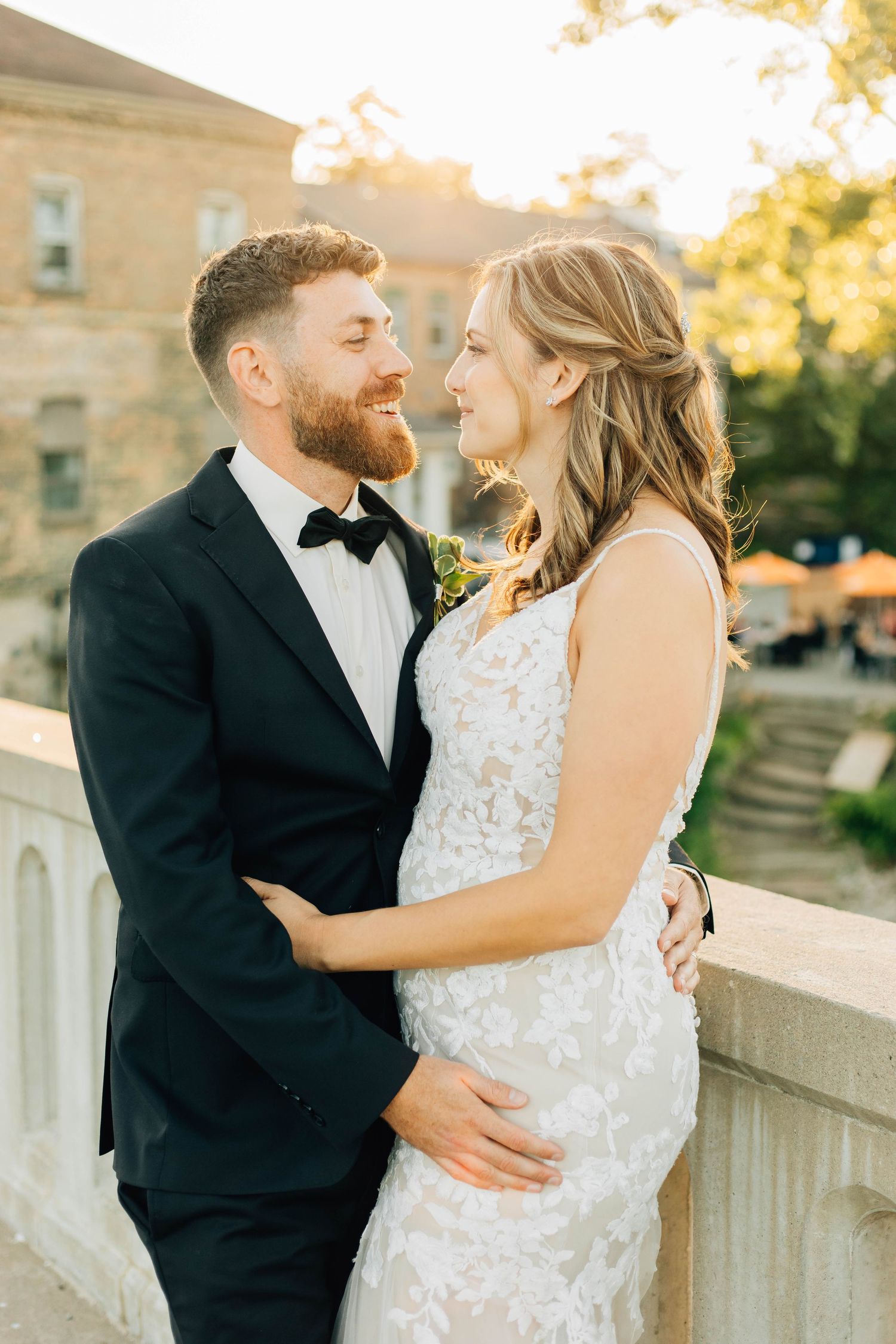 Smiling groom in black tuxedo and bride share a tender moment on a sunlit stone bridge in Paris Ontario.