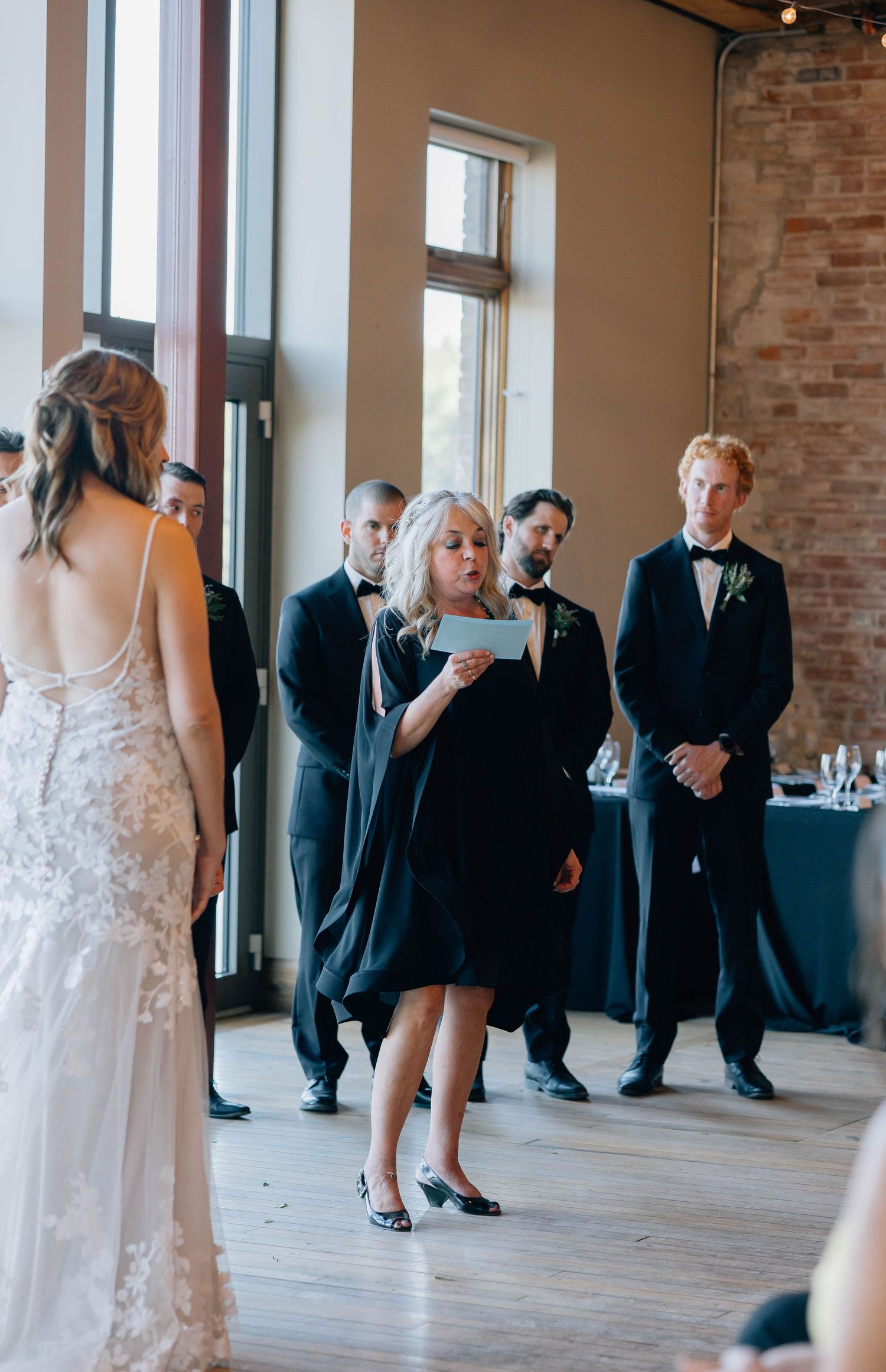 Woman in black dress reading from paper during wedding ceremony with groomsmen behind.