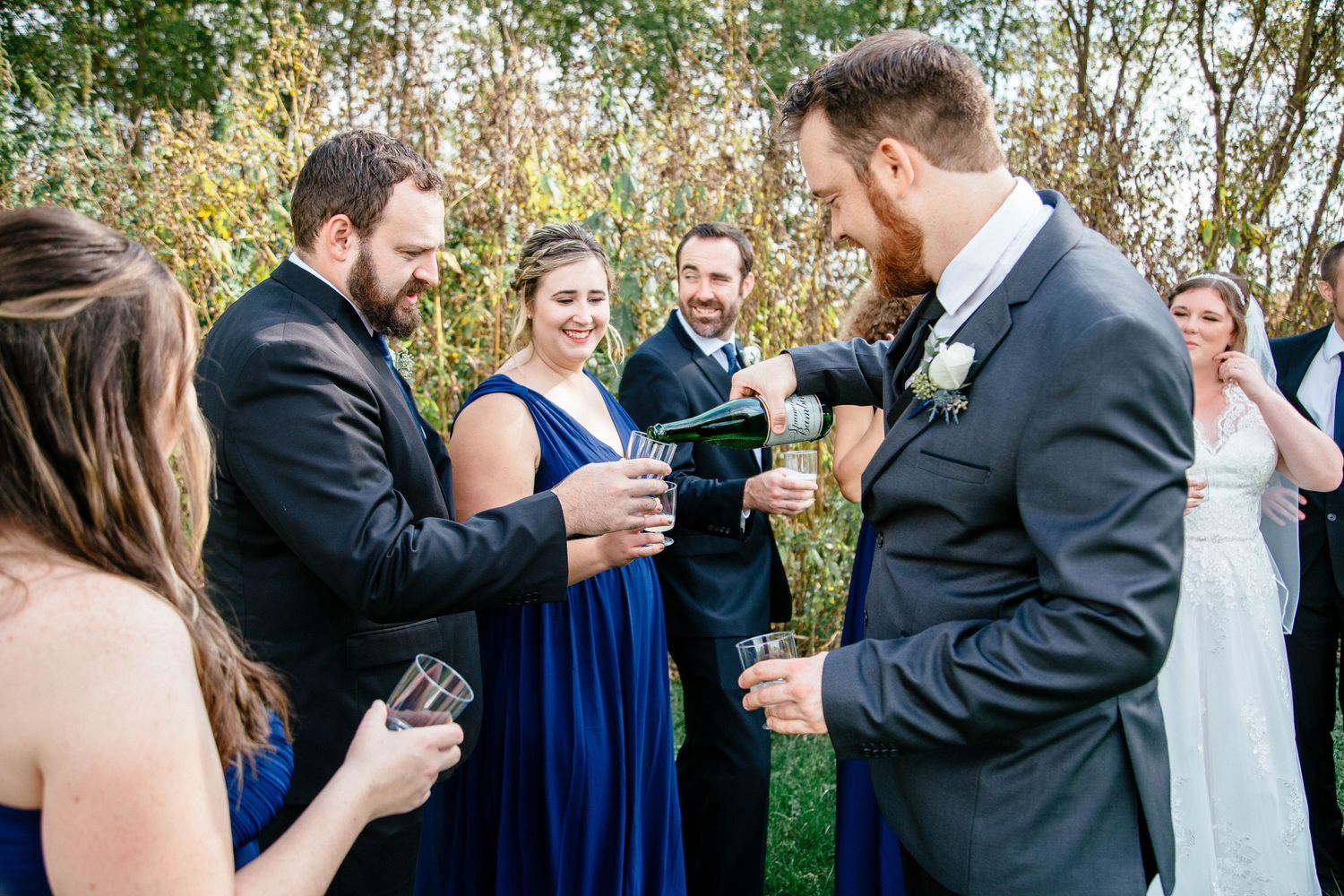 Groom pouring champagne for smiling wedding party guests outdoors during fall wedding celebration.