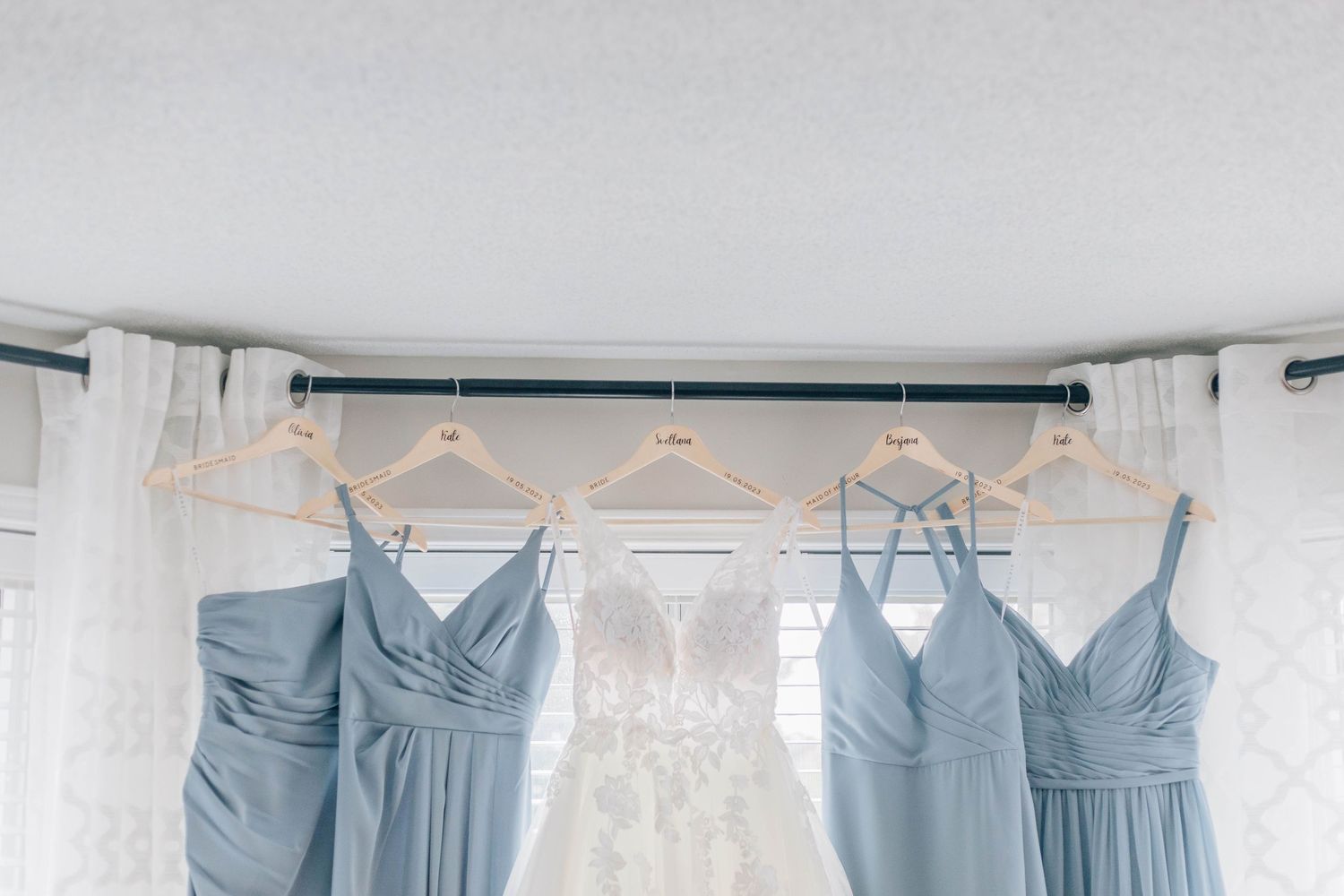 Dusty blue bridesmaid dresses and white wedding gown hanging on curtain rod near window.