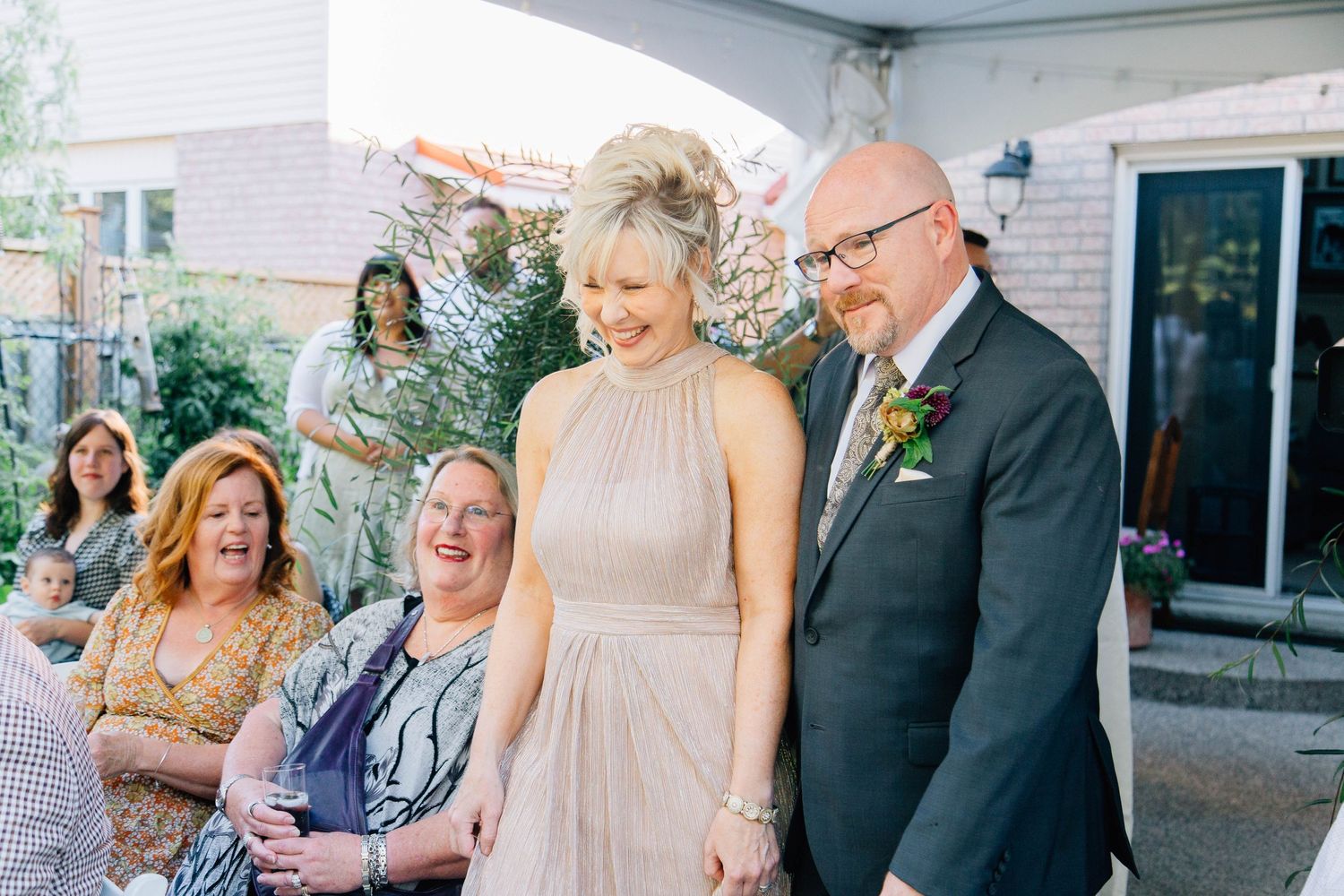 Elegantly dressed wedding guests laugh and celebrate together under a tent at an outdoor backyard wedding reception.