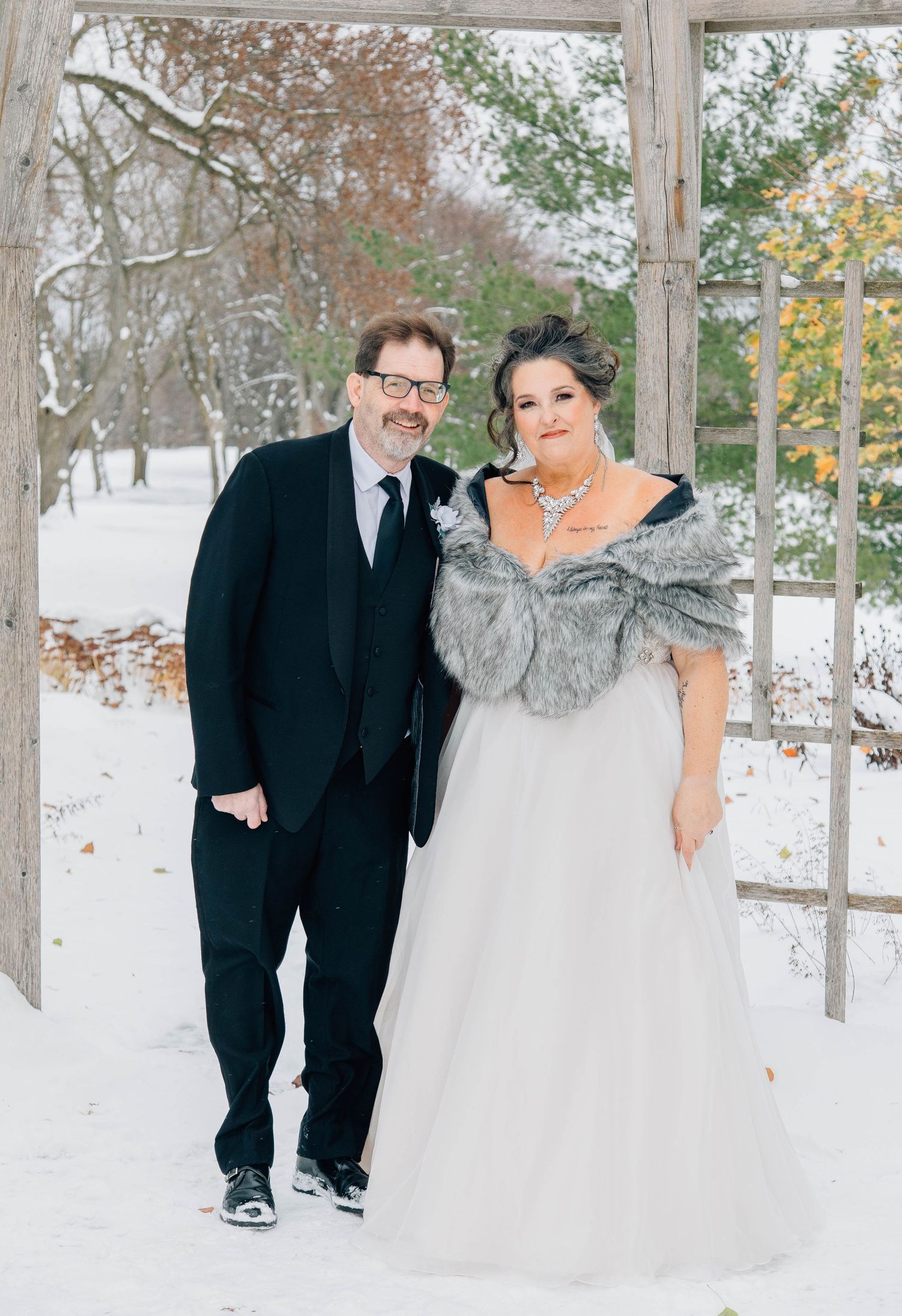 Bride and groom pose together in snowy winter wedding setting under rustic wooden arbor.
