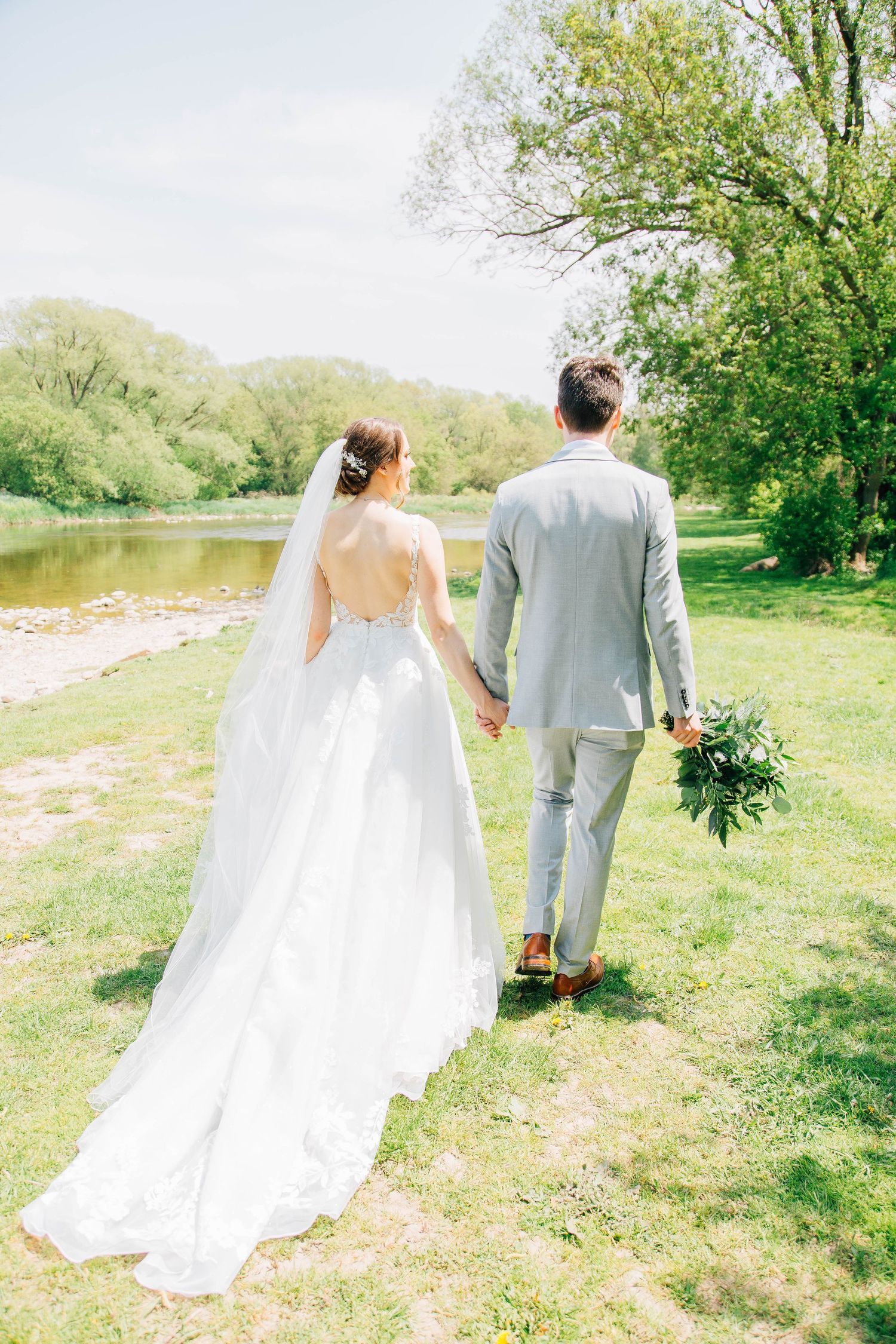 Bride and groom walk hand in hand toward a sunlit river on their wedding day.