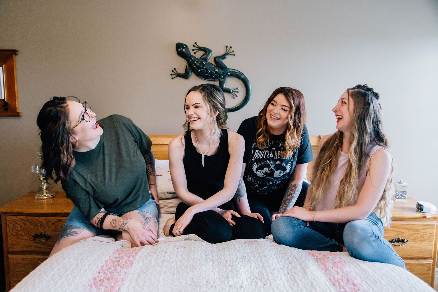 Four laughing women sitting casually on bed in cozy bedroom setting.