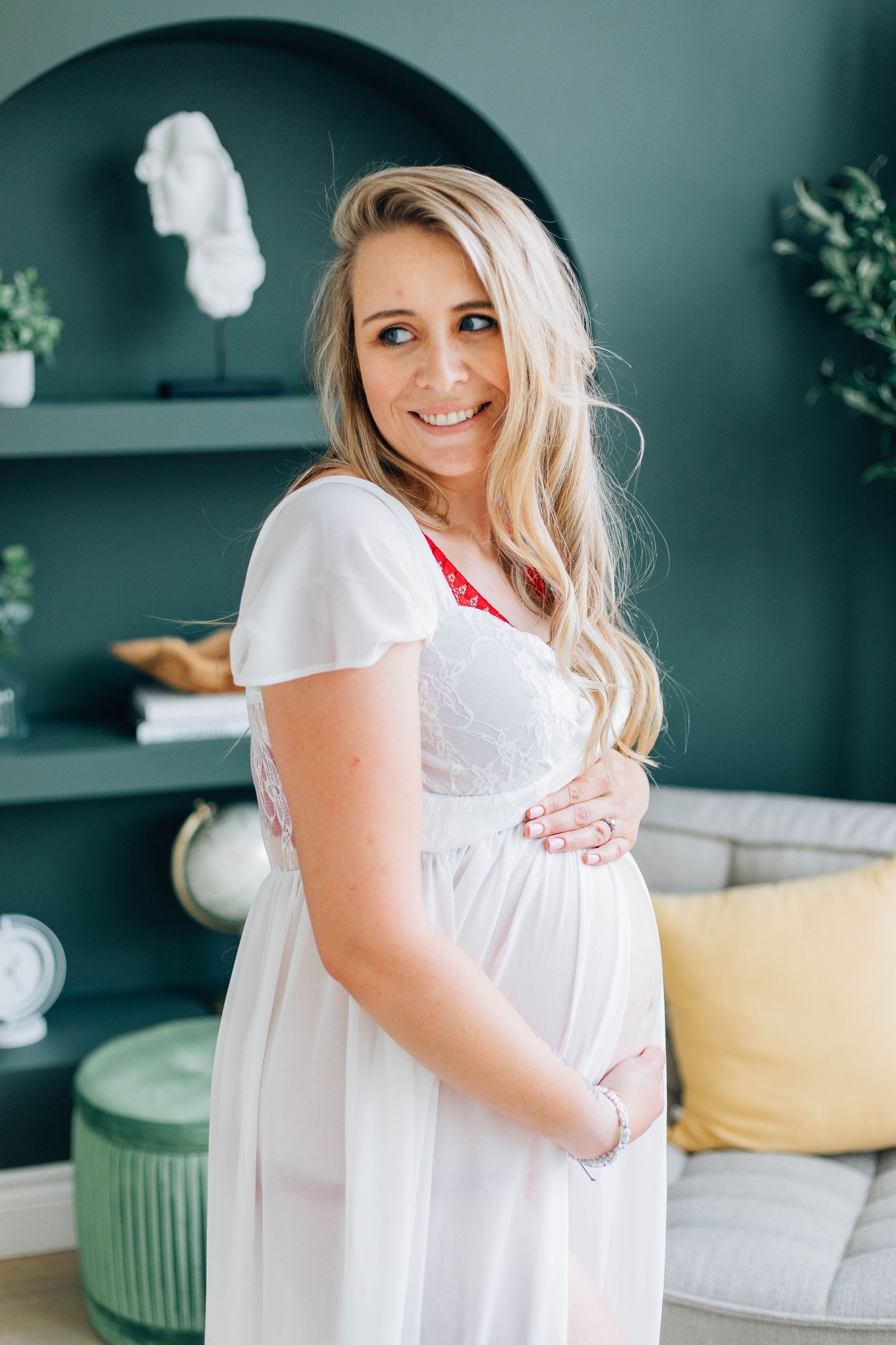 Smiling pregnant woman in white dress holding baby bump in styled room.
