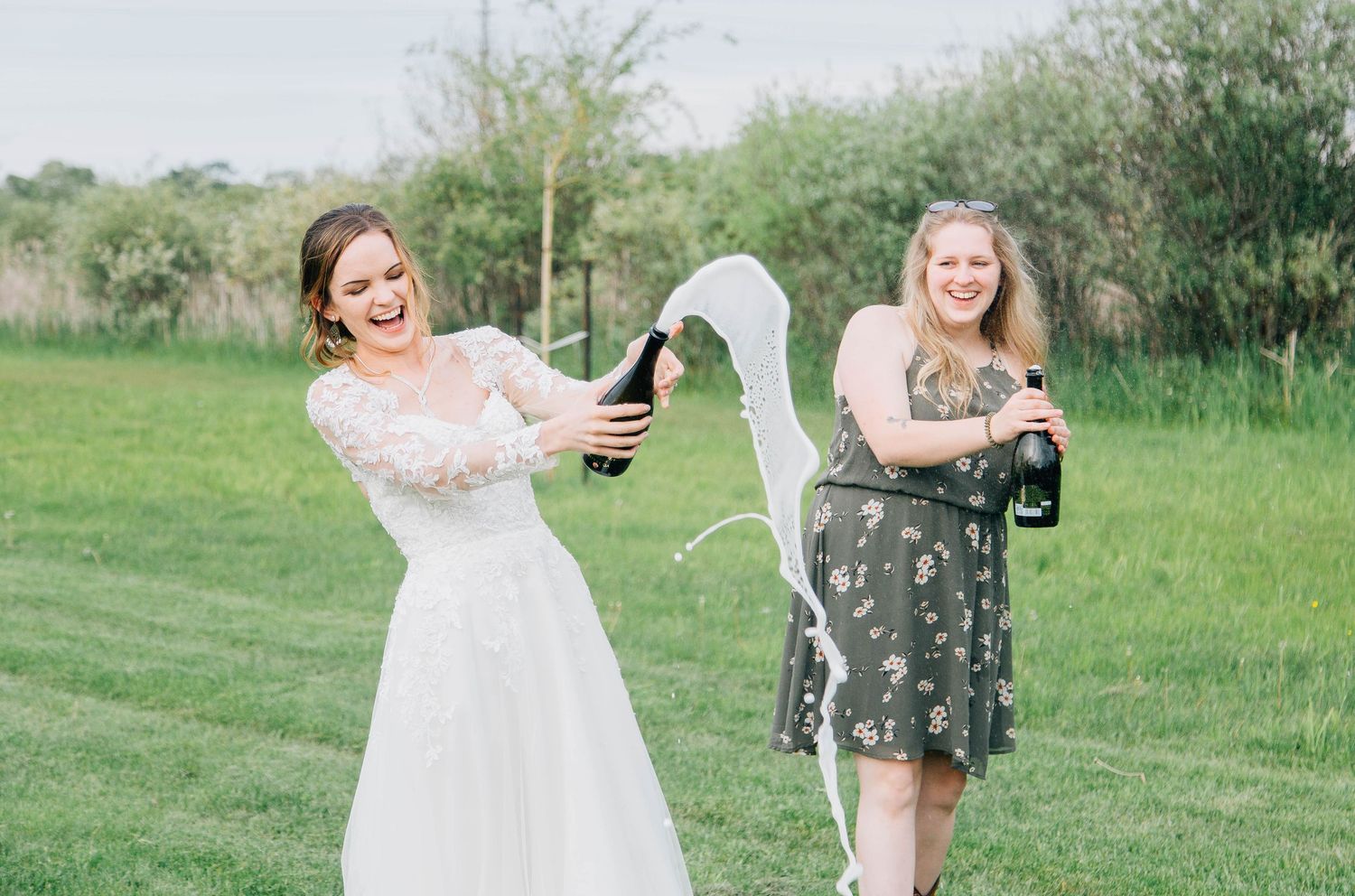 Joyful bride in white lace dress popping champagne with a friend at outdoor wedding.