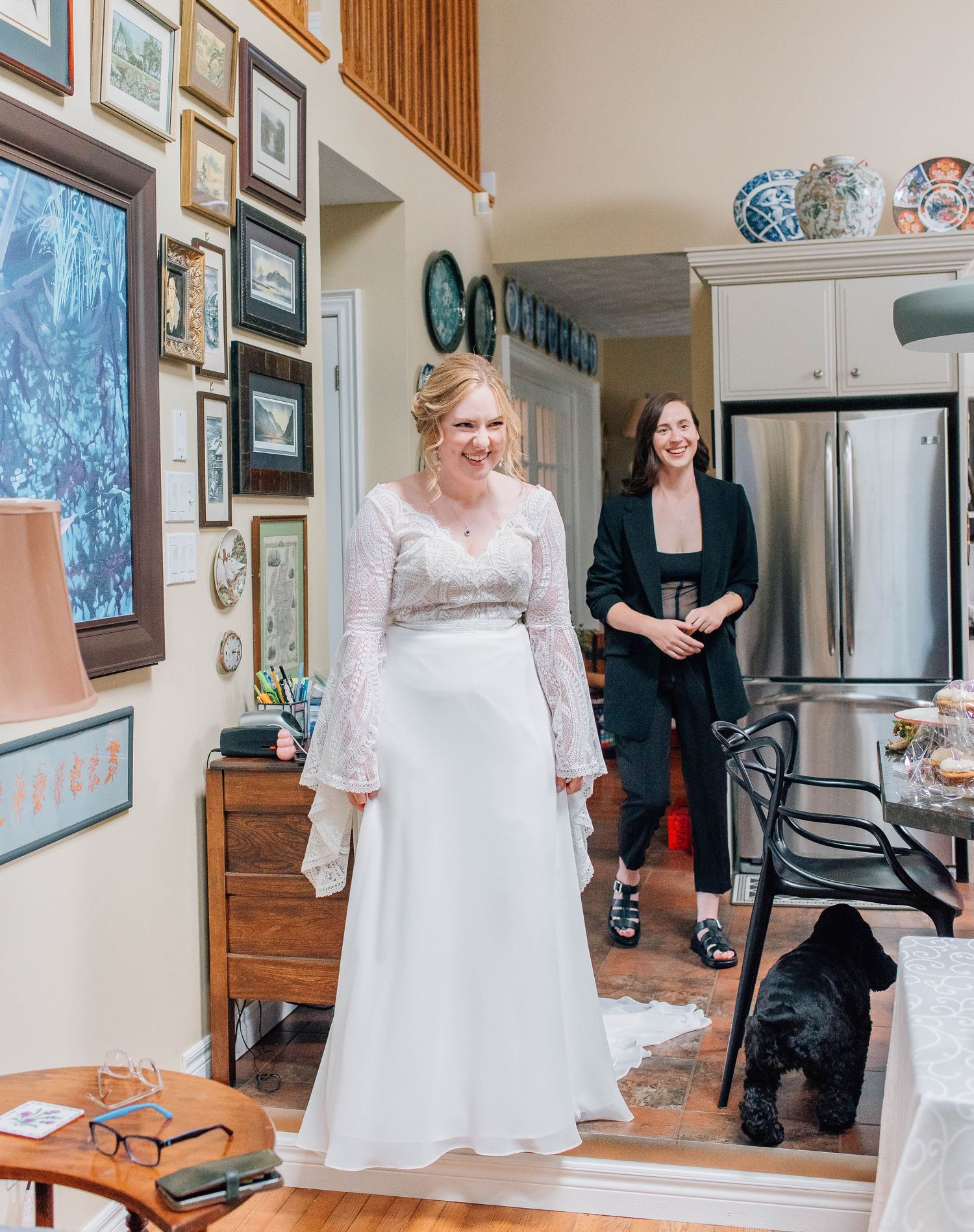 Bride in white lace dress smiles in decorated home hallway with black dog nearby.