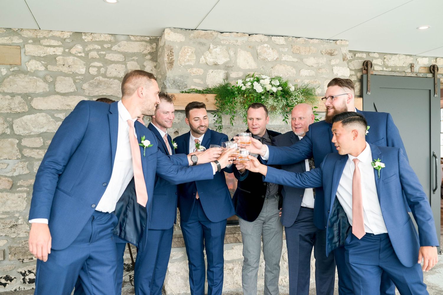 Groomsmen in navy suits with pink ties toast together in rustic stone wedding venue.