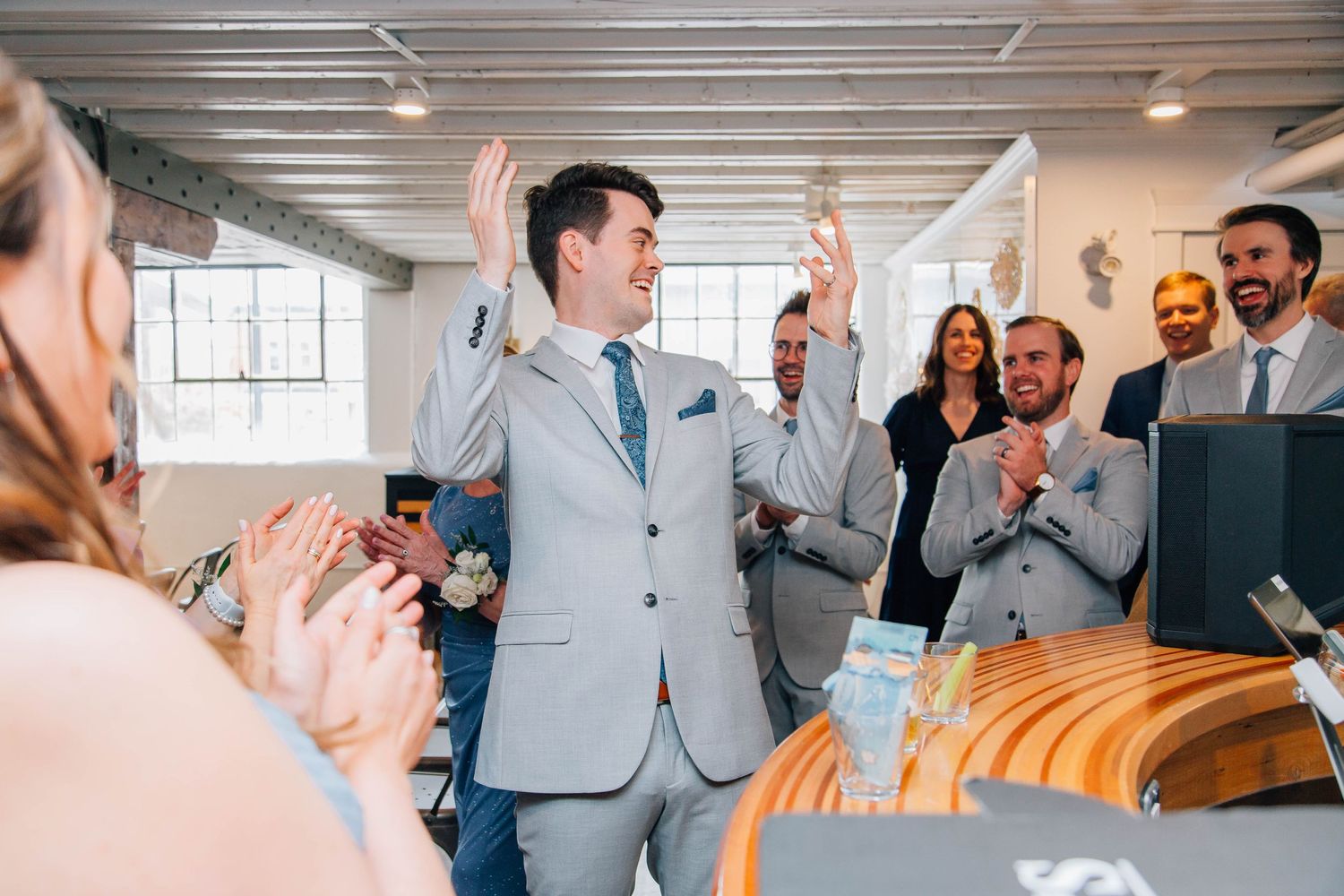 Excited groom raises hands triumphantly as wedding guests applaud and celebrate at indoor reception venue.