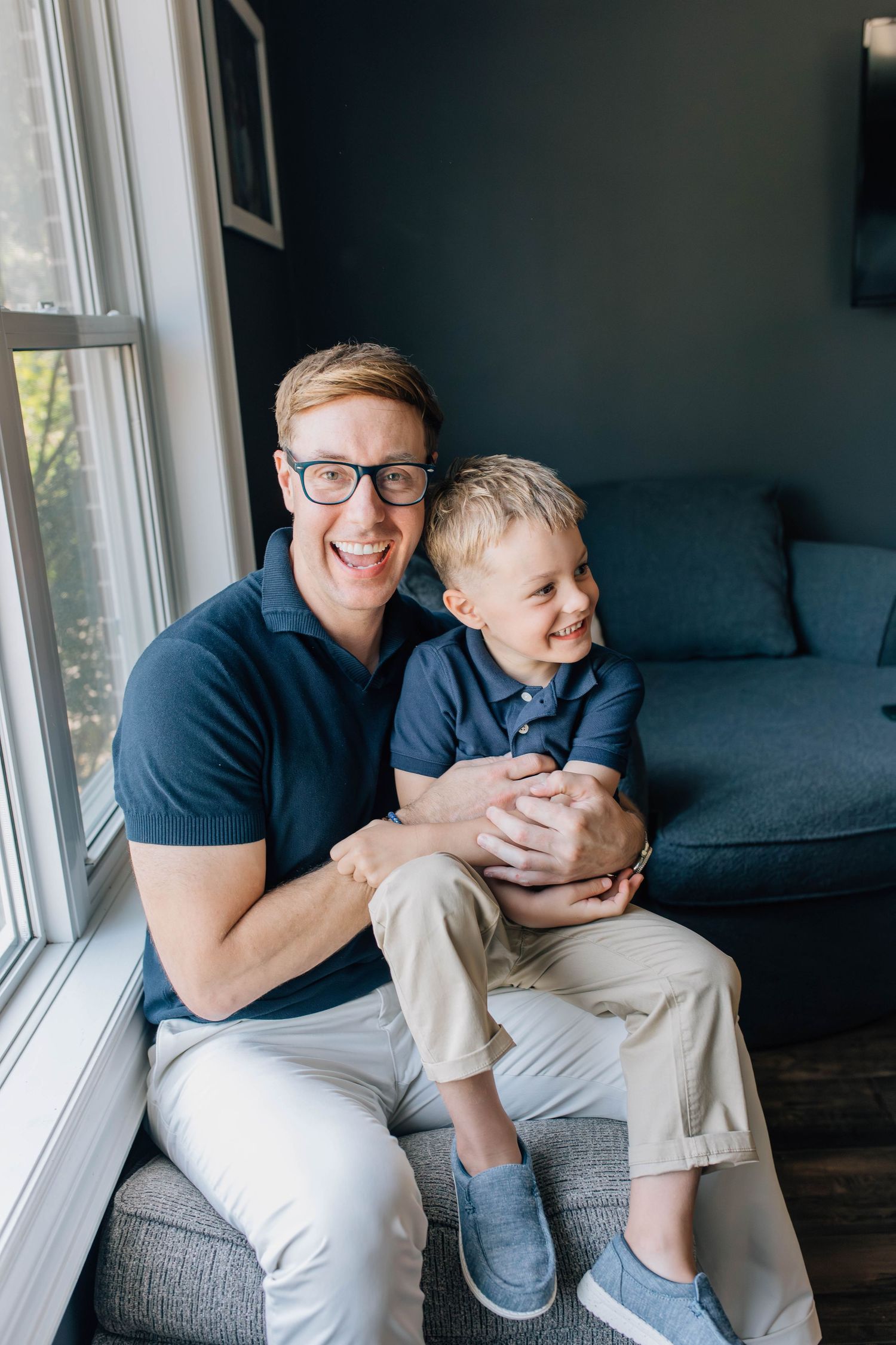Laughing dad with glasses sitting by window holding smiling toddler son in matching navy polo shirts.