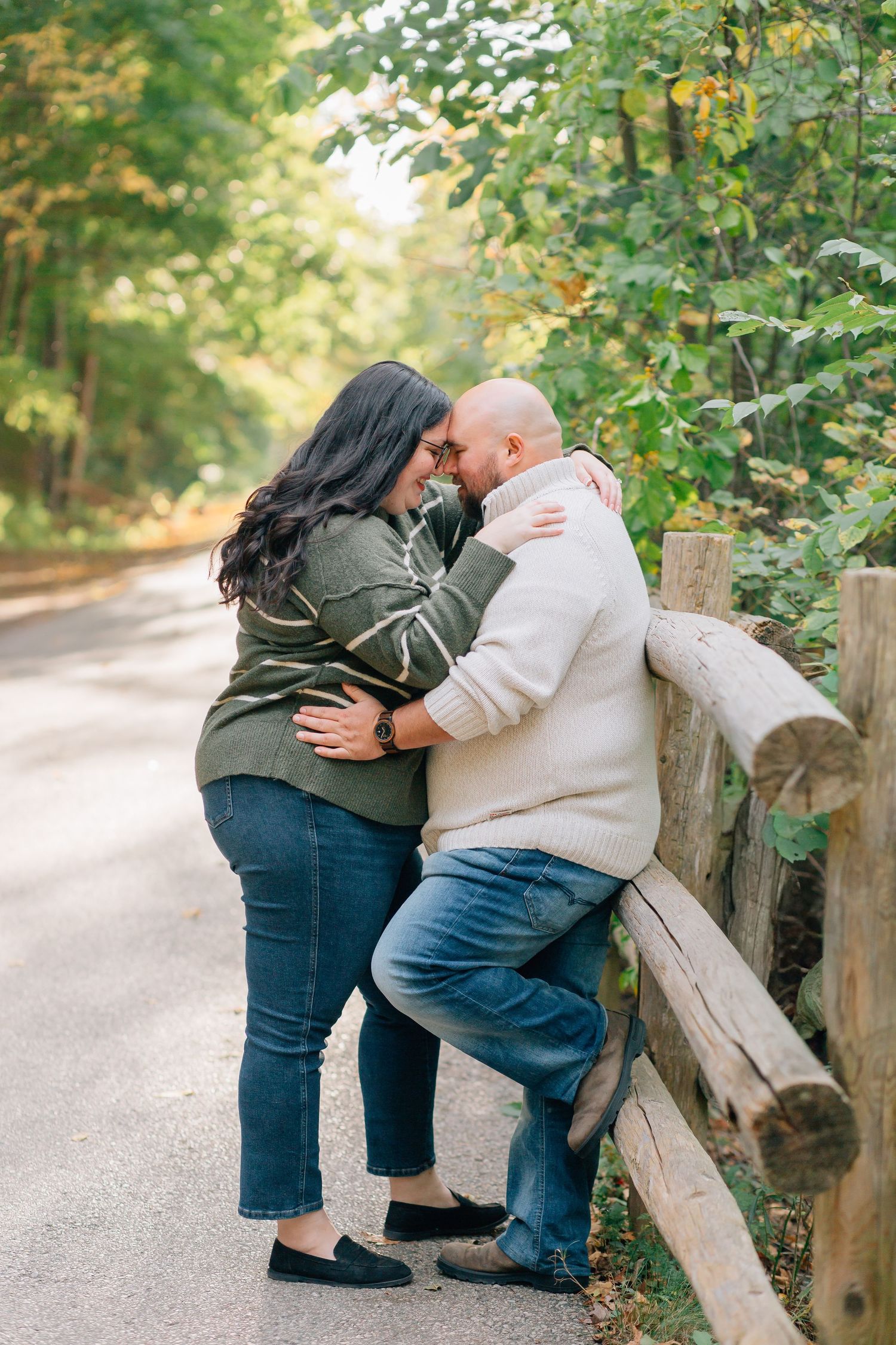 Couple embracing and touching foreheads by a rustic wooden fence on a tree-lined road.