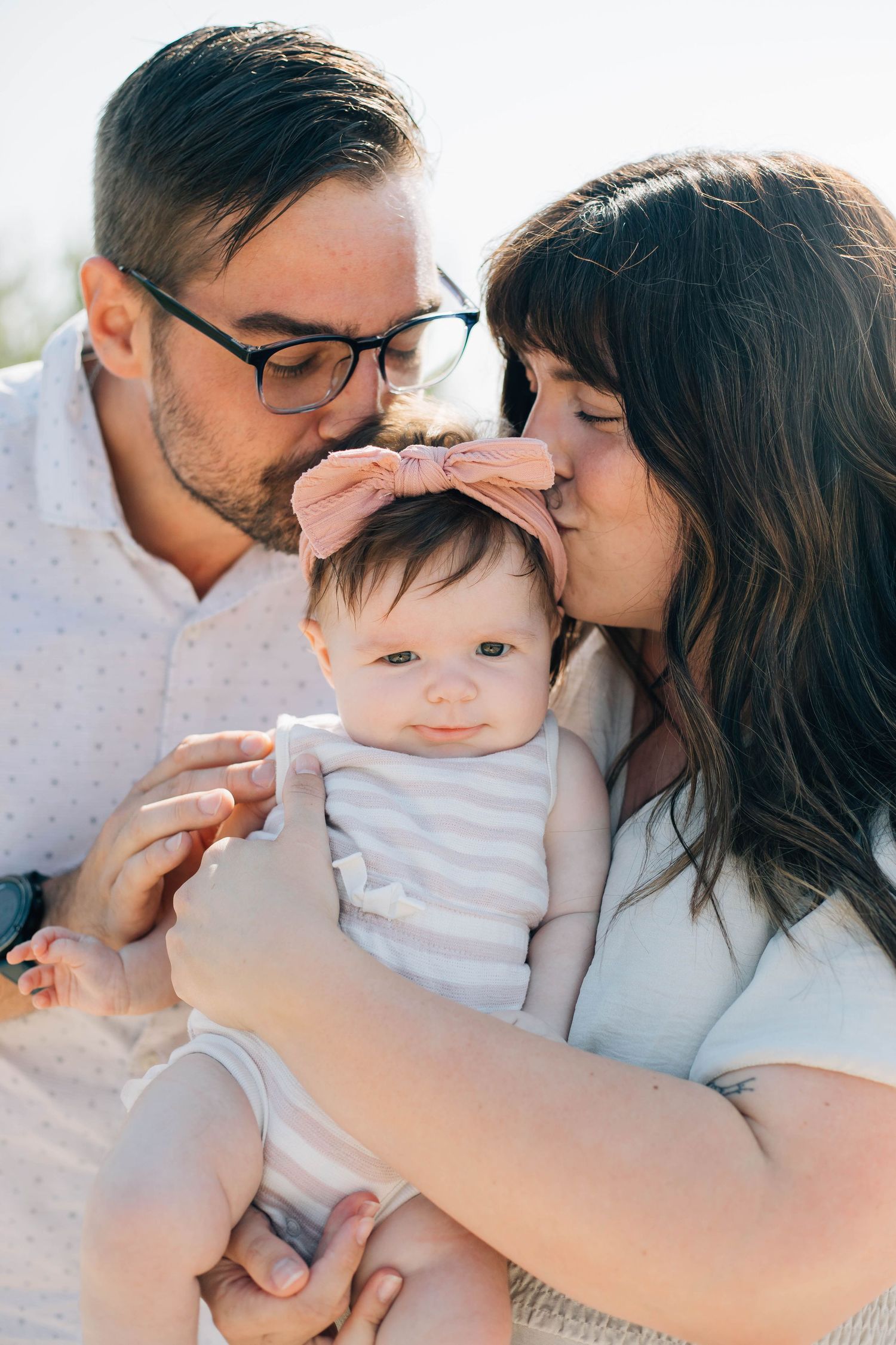 Parents kissing their baby girl wearing a pink bow headband during an outdoor family portrait session.