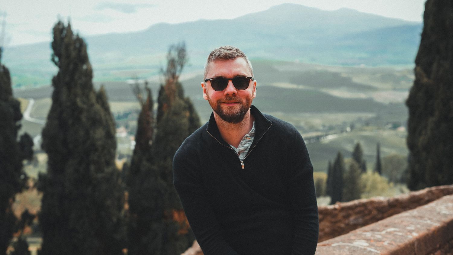 A person wearing sunglasses and a black sweater sits on a stone wall with cypress trees and rolling hills in the background.