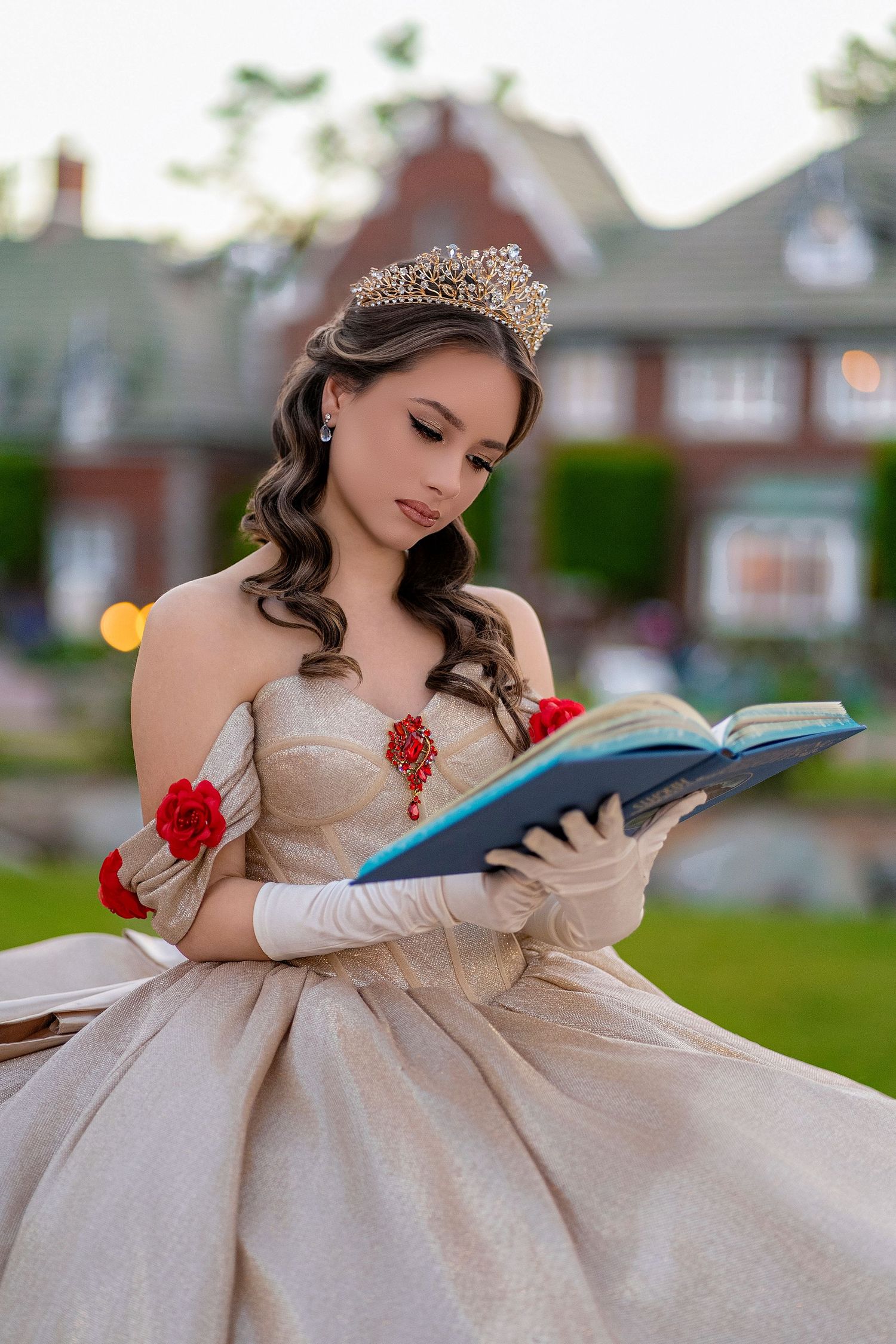Young woman in champagne ball gown with tiara and gloves reads book in garden setting.