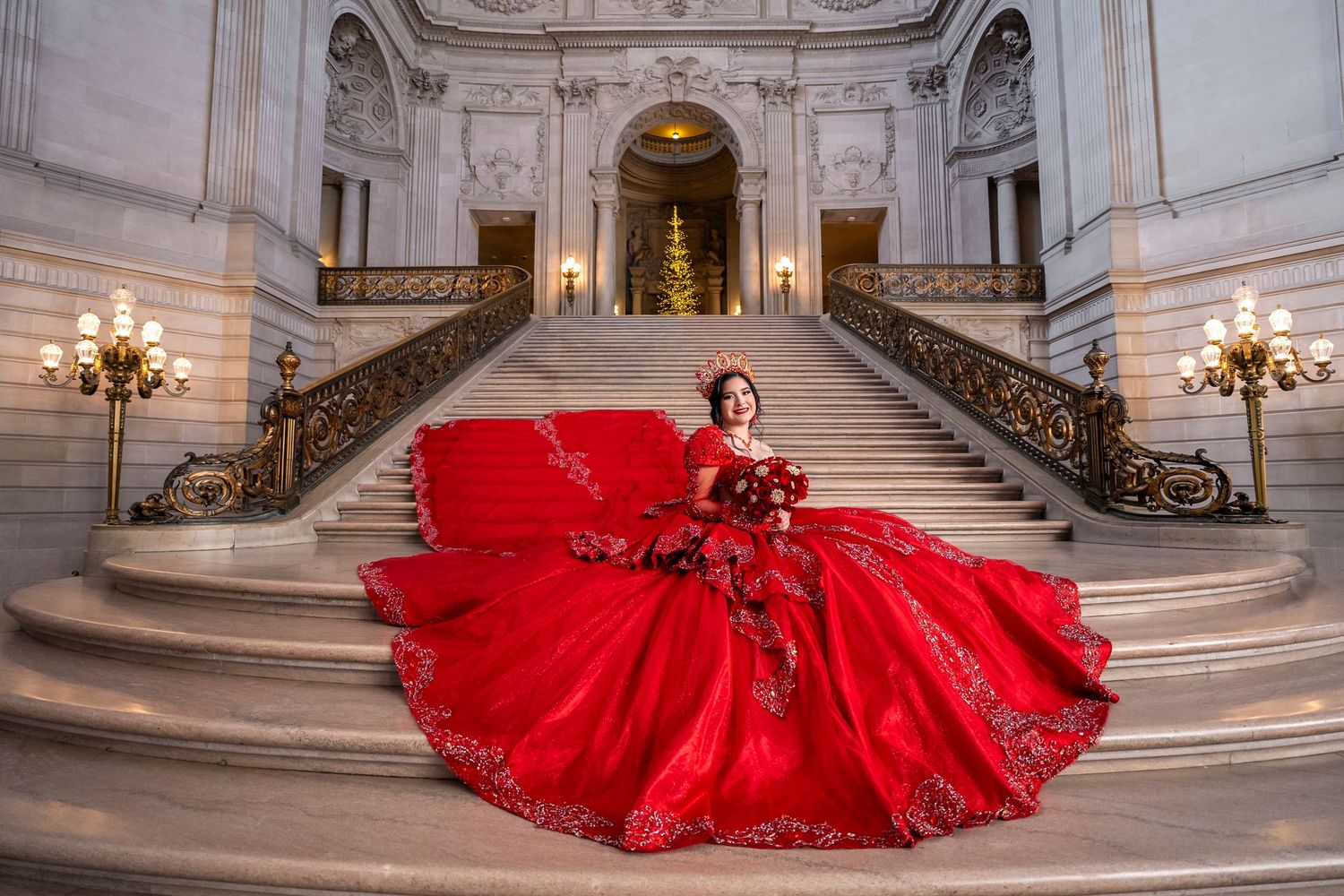 Woman in stunning red ballgown poses gracefully on marble staircase in elegant grand hall with holiday decor.