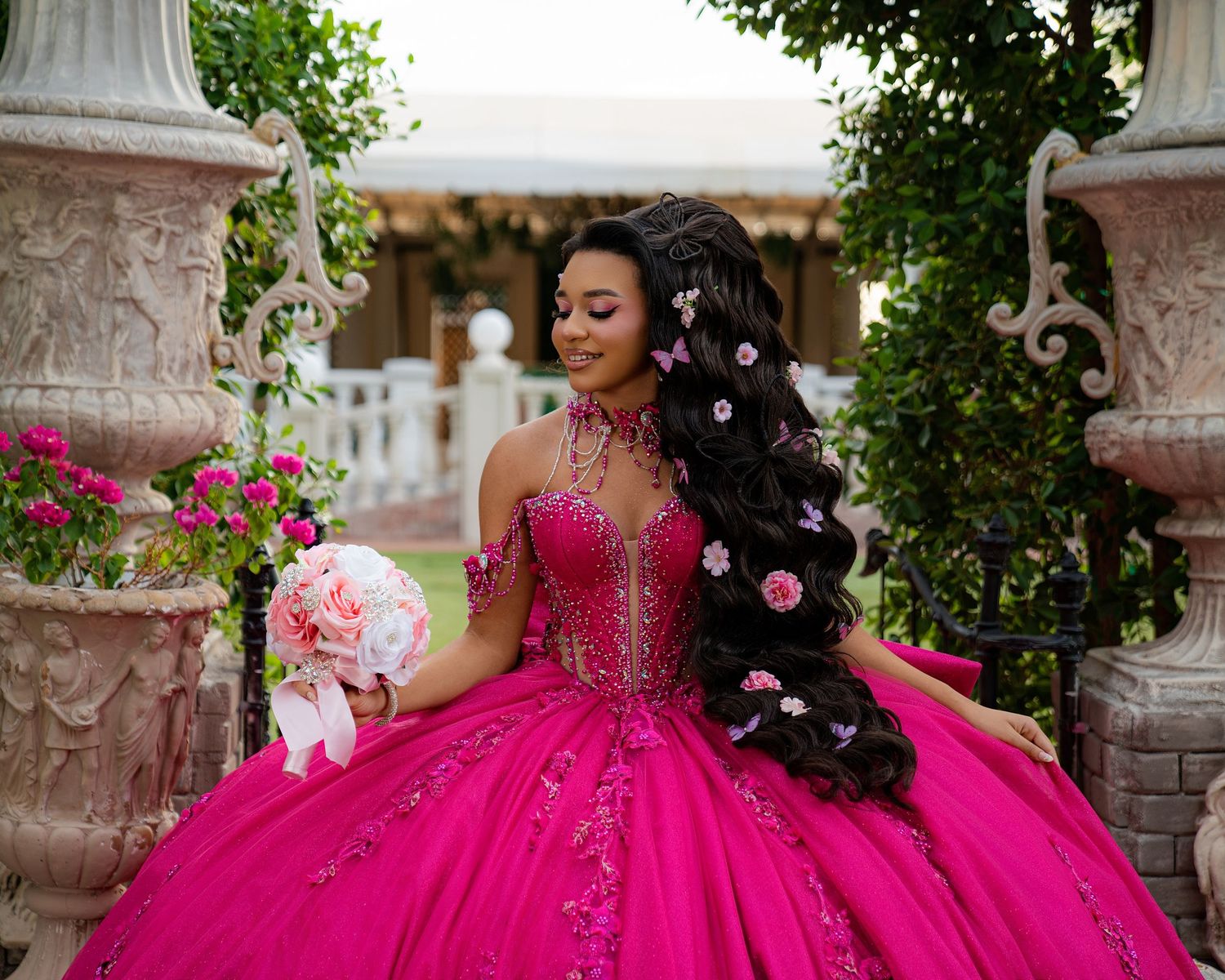 Young woman in a stunning fuchsia ball gown with floral hair accessories holds a bouquet at an elegant outdoor venue.