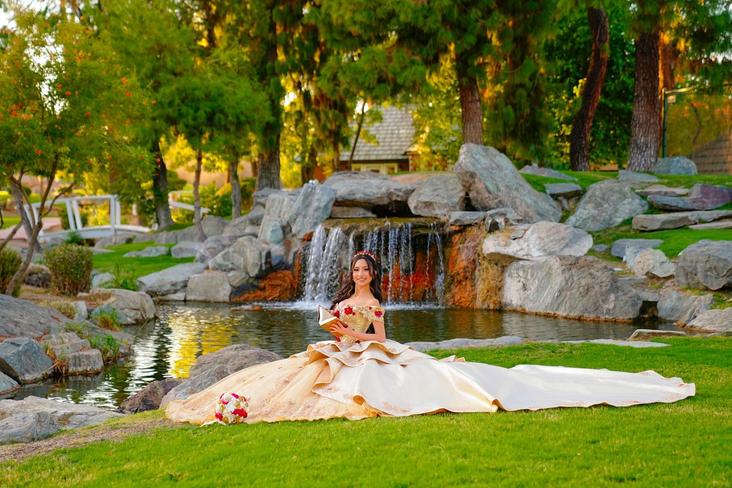 Young woman in a dramatic gold quinceañera ball gown with long train seated by a garden waterfall.