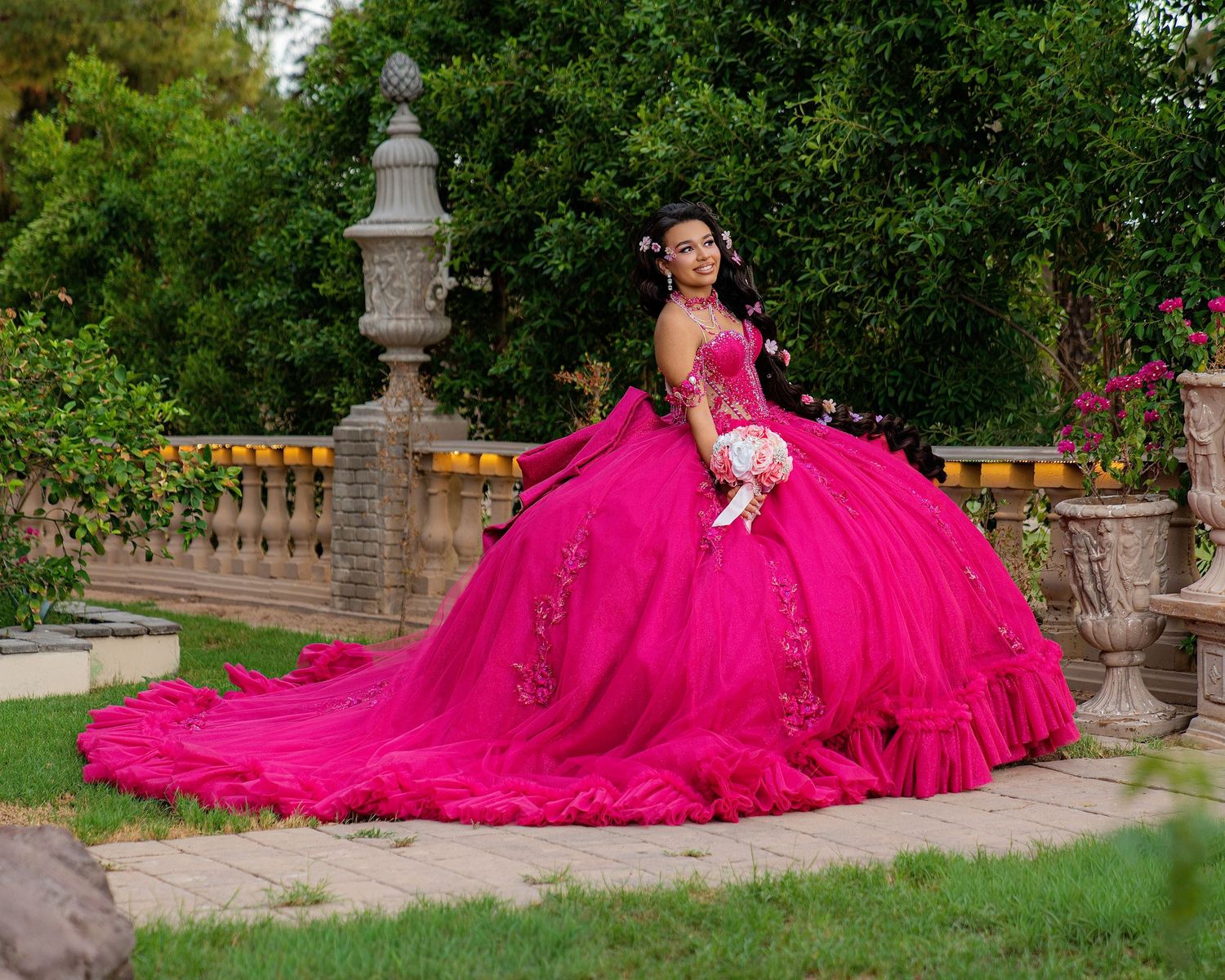Young woman in a vibrant fuchsia quinceañera ball gown with floral embroidery poses in a lush garden setting.