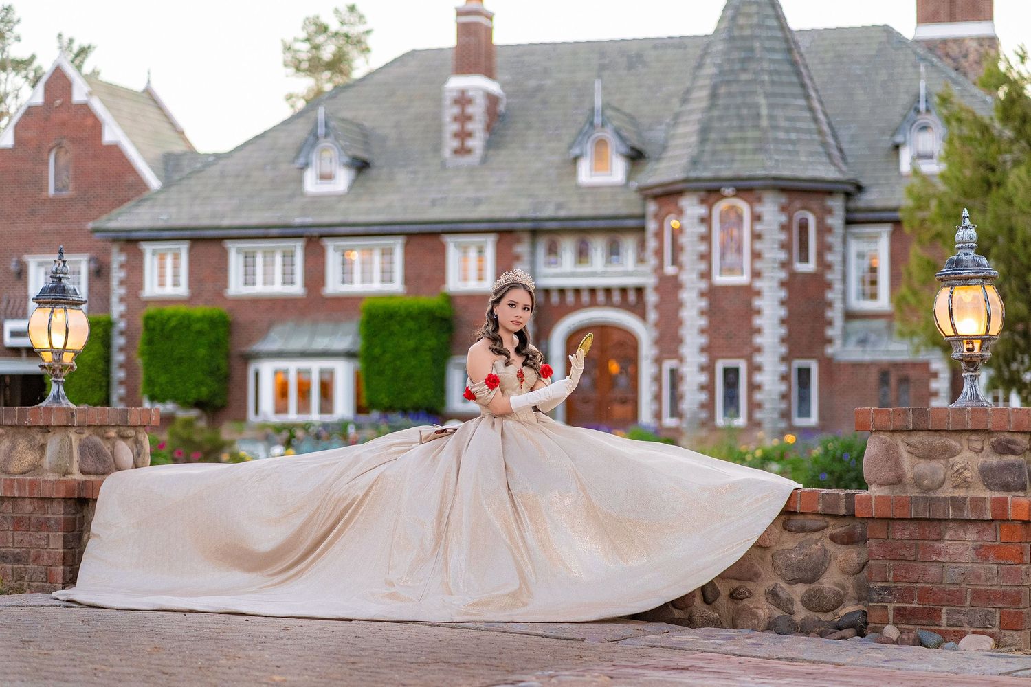 Young woman in elegant champagne ballgown twirling in front of a grand brick mansion at dusk.