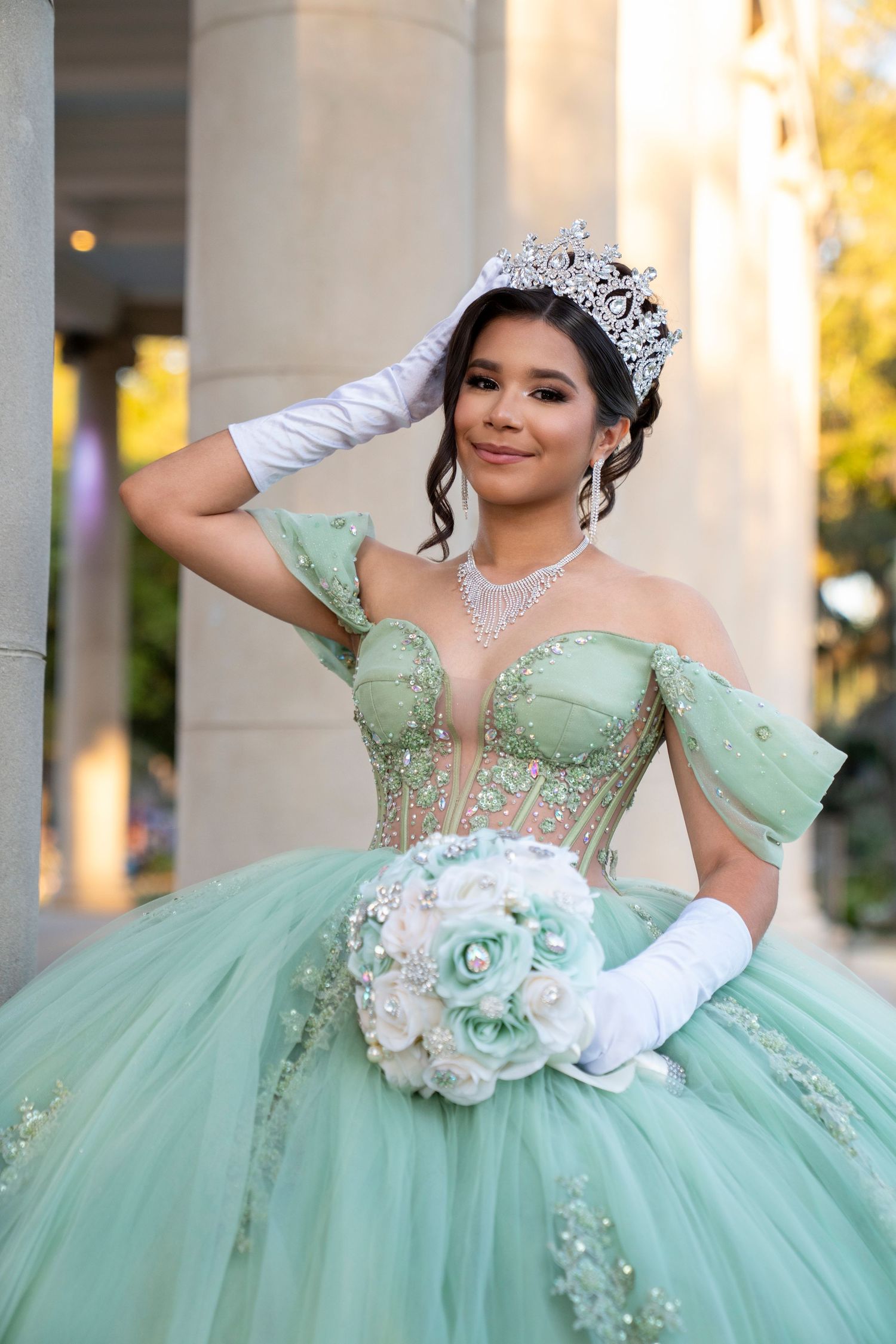 Young woman in mint green quinceañera ball gown with tiara, white gloves, and floral bouquet posing outdoors.