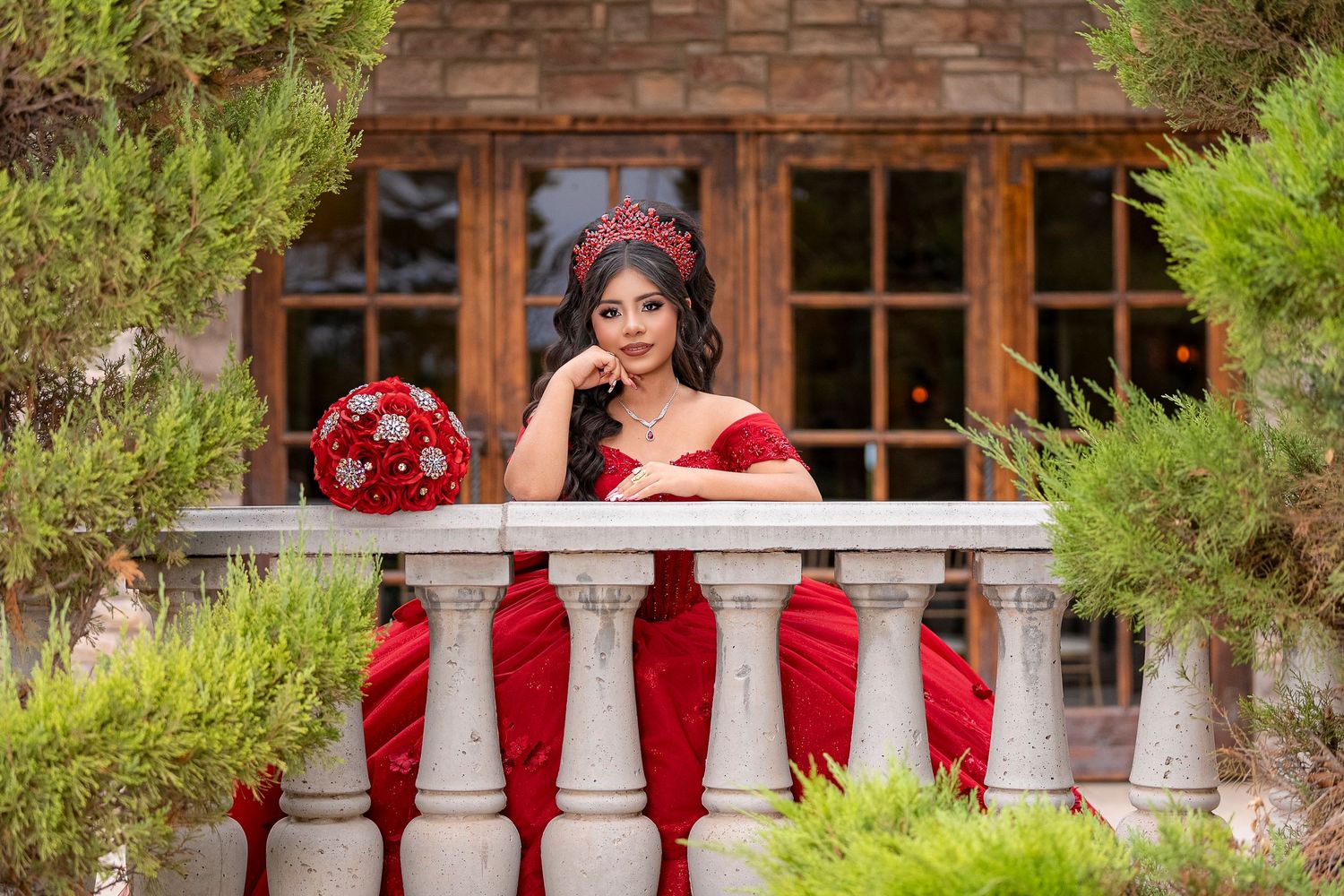 Young woman in red off-shoulder gown and crown holds red bouquet on stone balcony surrounded by lush greenery.