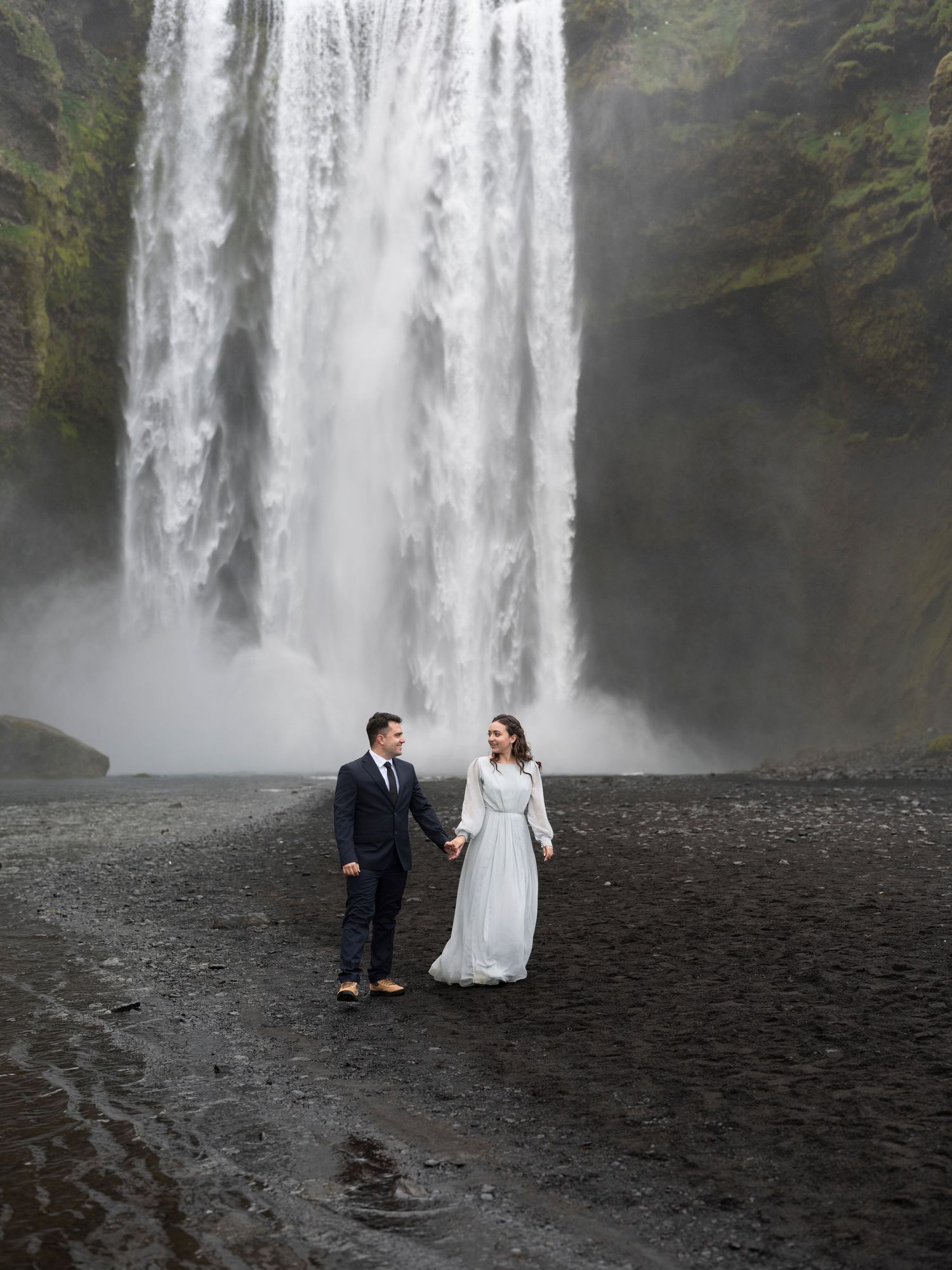 Couple in wedding attire holds hands before the massive Skógafoss waterfall in Iceland on a misty day.