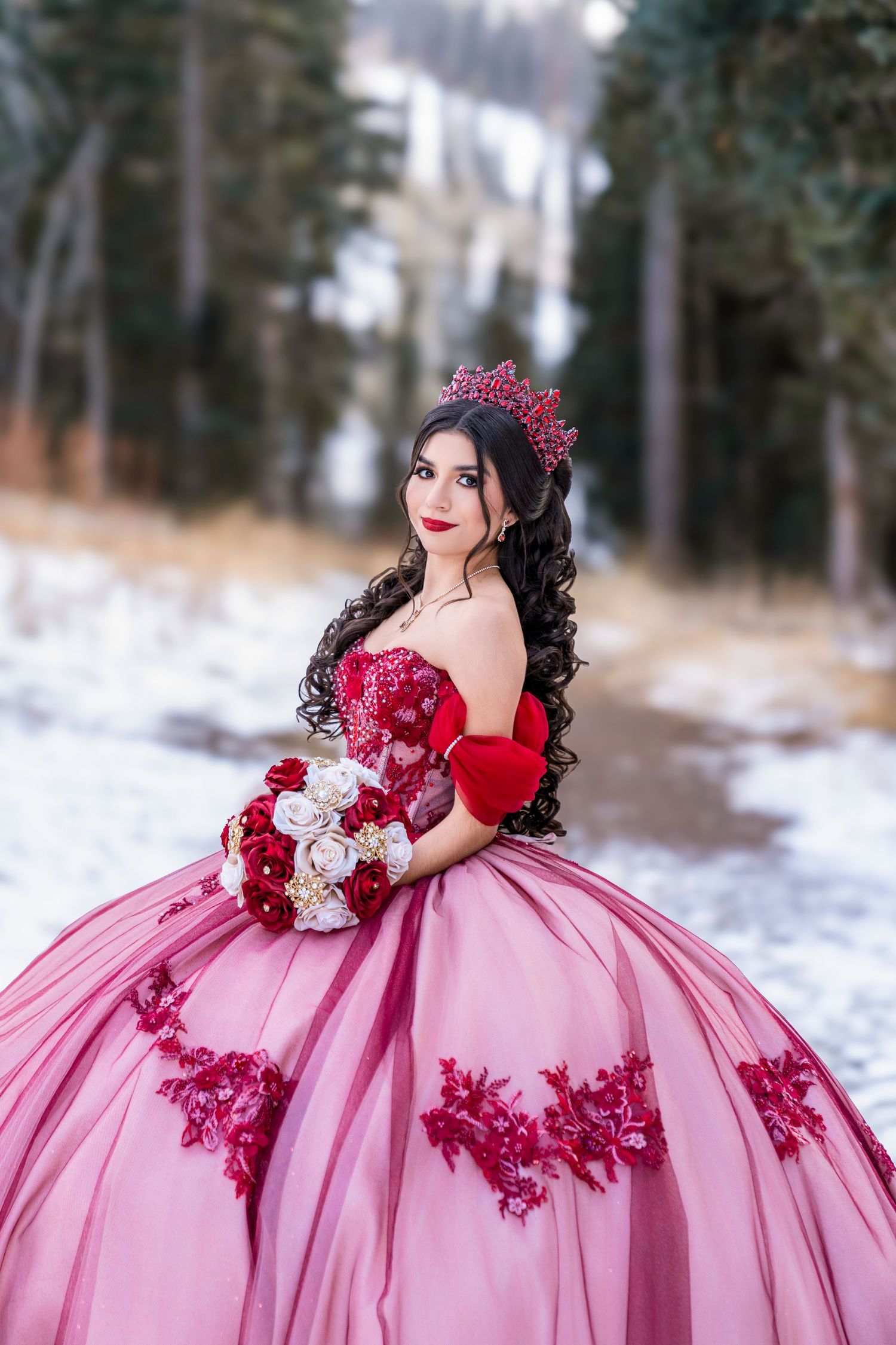 Young woman in a pink and red quinceañera ball gown holds a floral bouquet in a snowy winter forest.