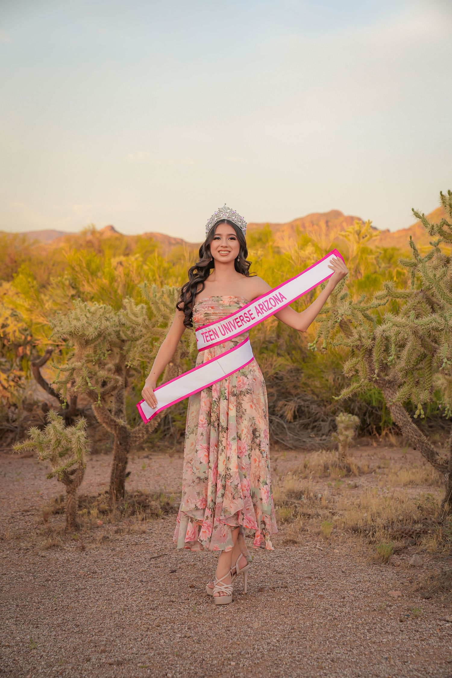 Young woman in floral gown wearing Miss Universe Arizona sash and crown poses in desert landscape with cacti.