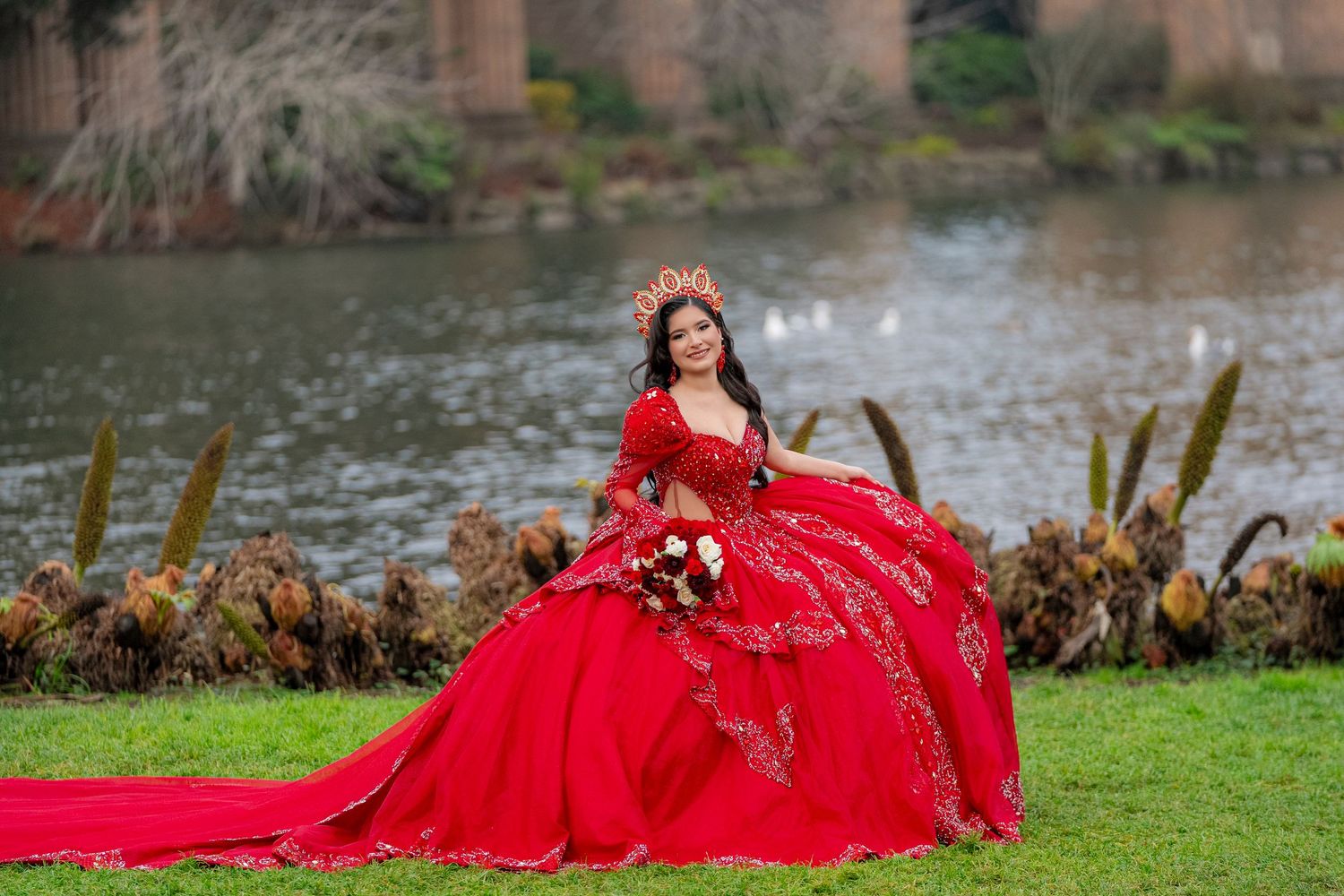 Young woman in elegant red quinceañera ball gown with floral crown holds bouquet beside a serene lake.