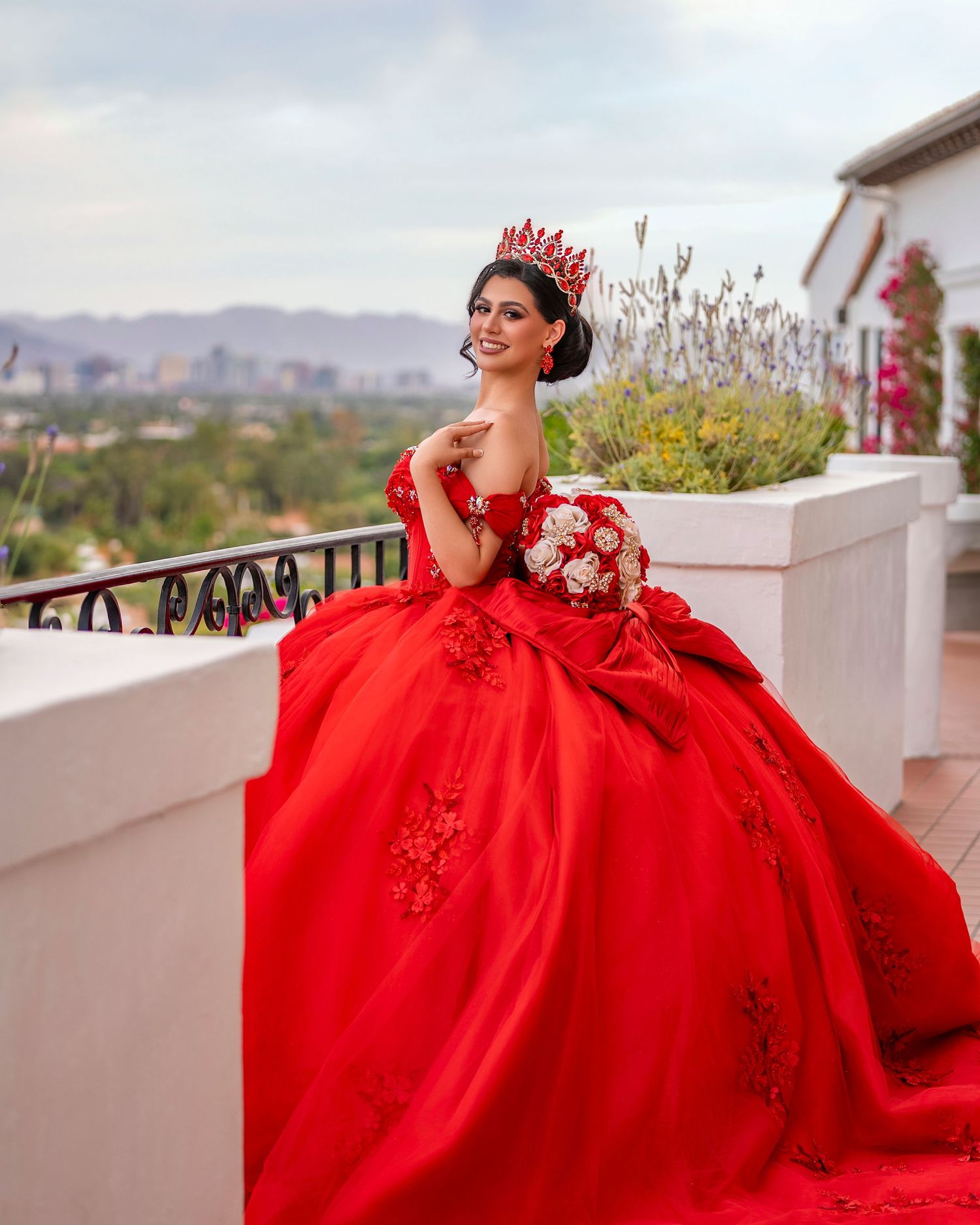Young woman in a stunning red ball gown with crown and bouquet poses on a balcony for quinceañera.