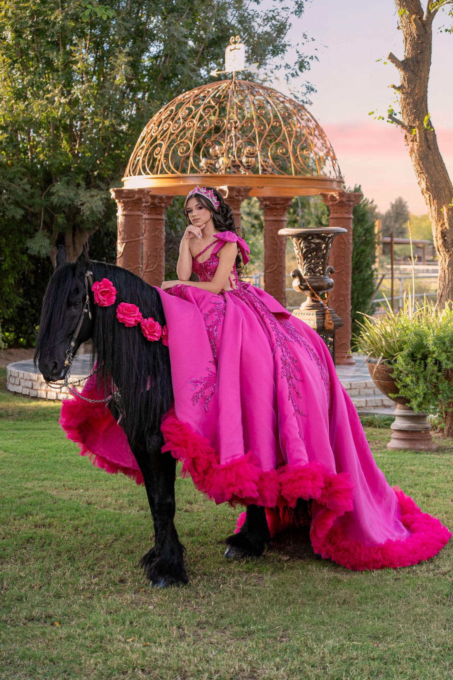 Young woman in pink quinceañera gown rides black horse near garden gazebo.