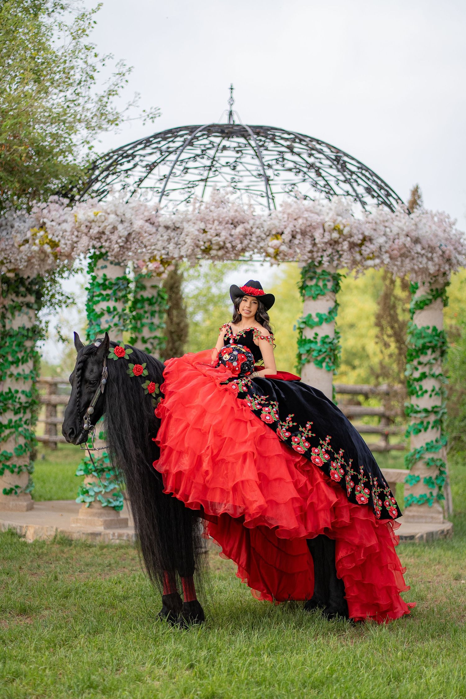 Woman in red and black ball gown riding a black horse under a floral gazebo outdoors.