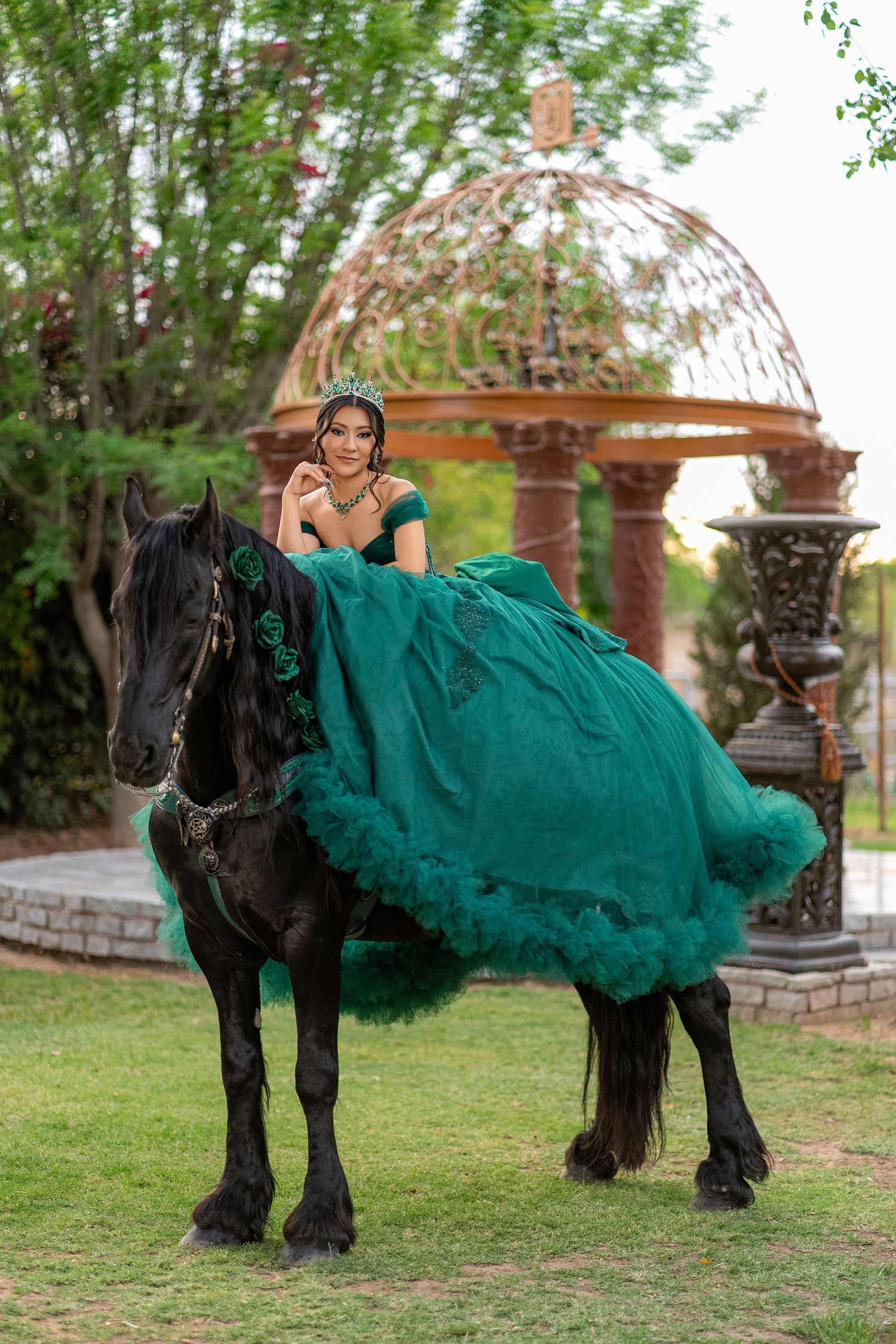 Young woman in emerald green quinceañera gown seated on black horse near ornate gazebo in garden.
