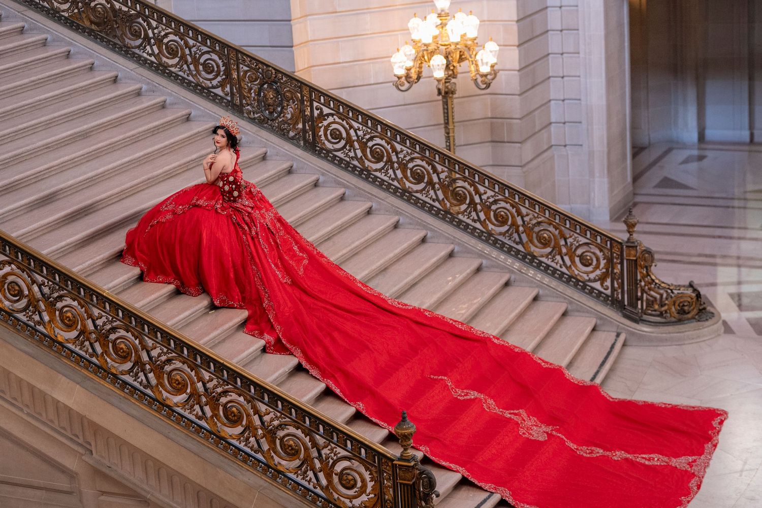 Woman in stunning red ballgown with long train posed on ornate grand staircase in elegant classical building.