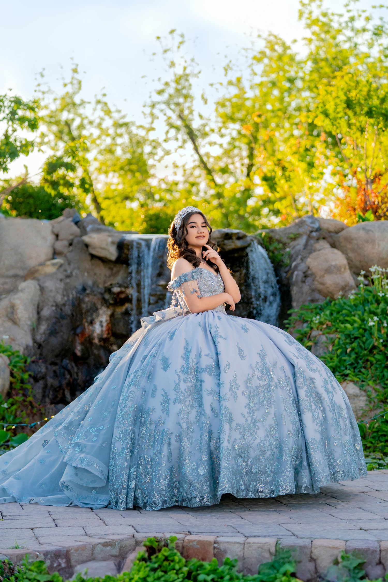Young woman in a light blue ball gown quinceañera dress poses near a waterfall surrounded by lush greenery.
