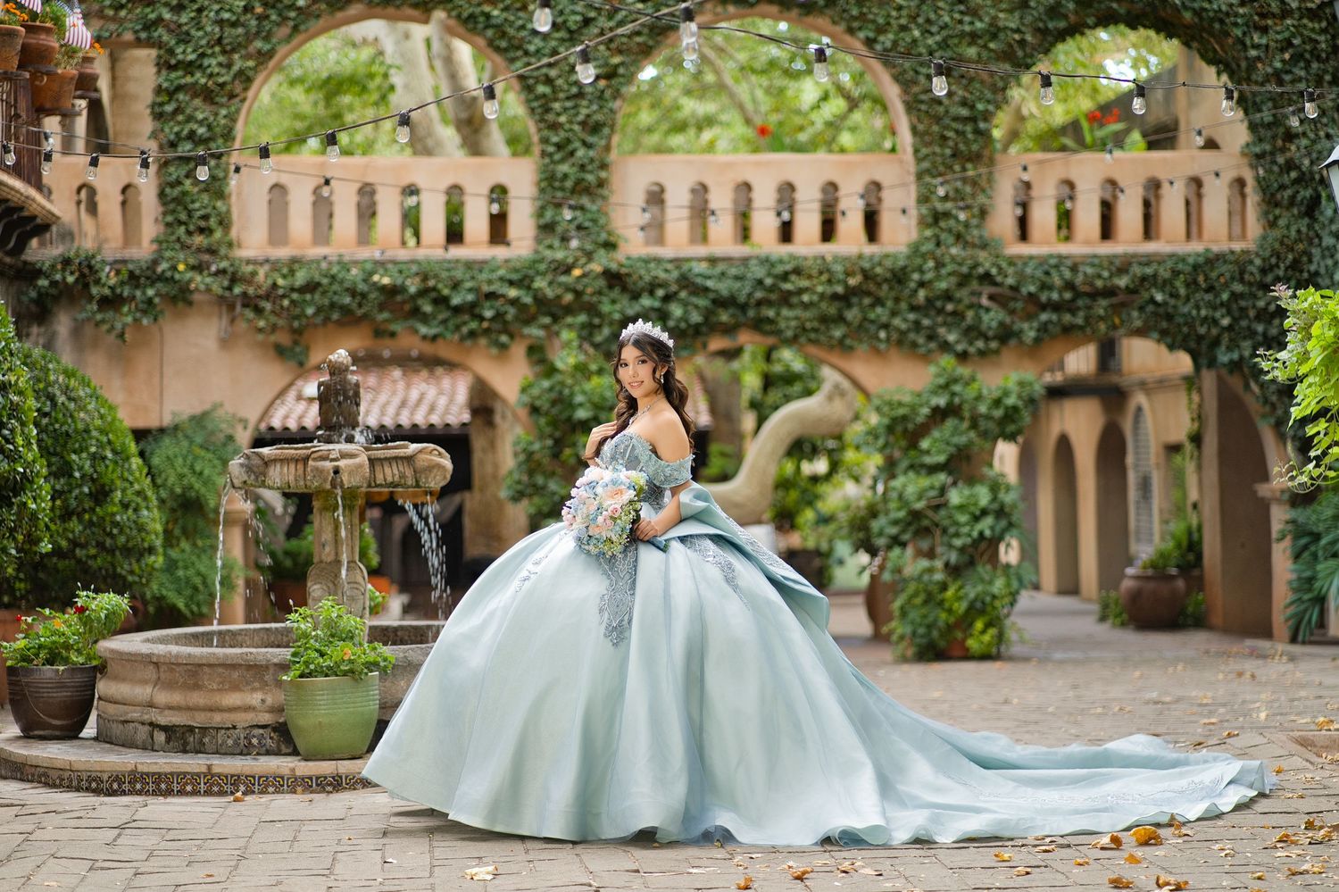 A young woman in a light blue ball gown quinceañera dress poses in a lush courtyard with ivy-covered arches.