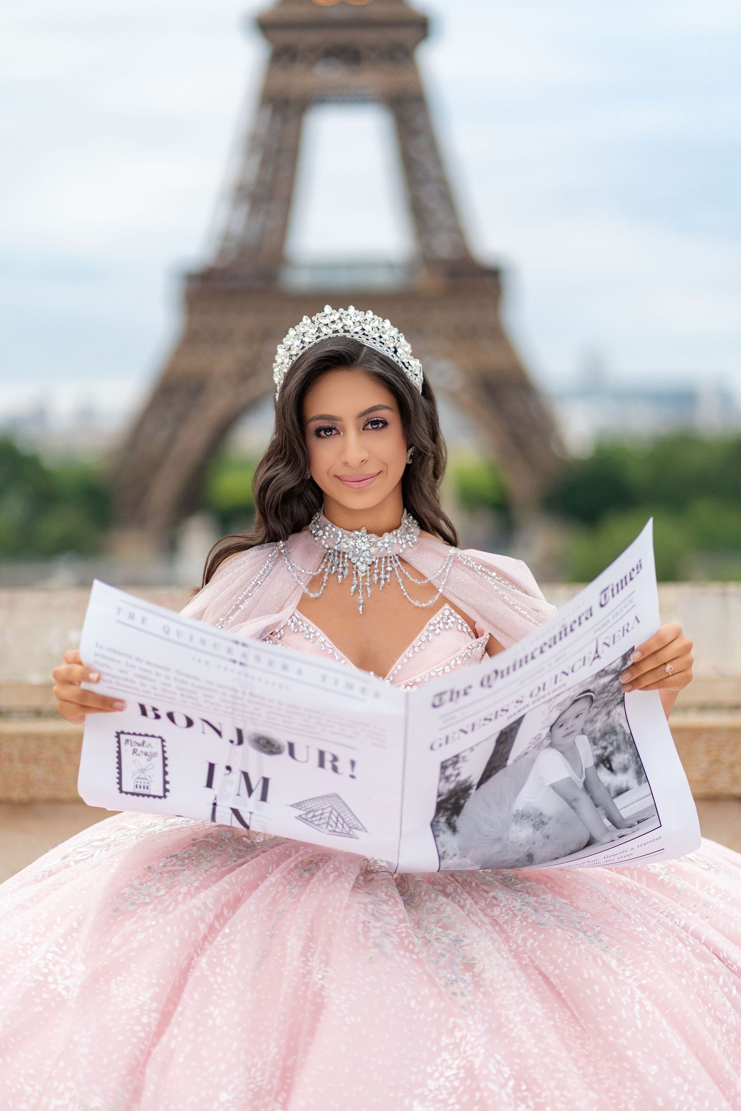 Young woman in pink quinceañera dress reads newspaper before the Eiffel Tower in Paris.