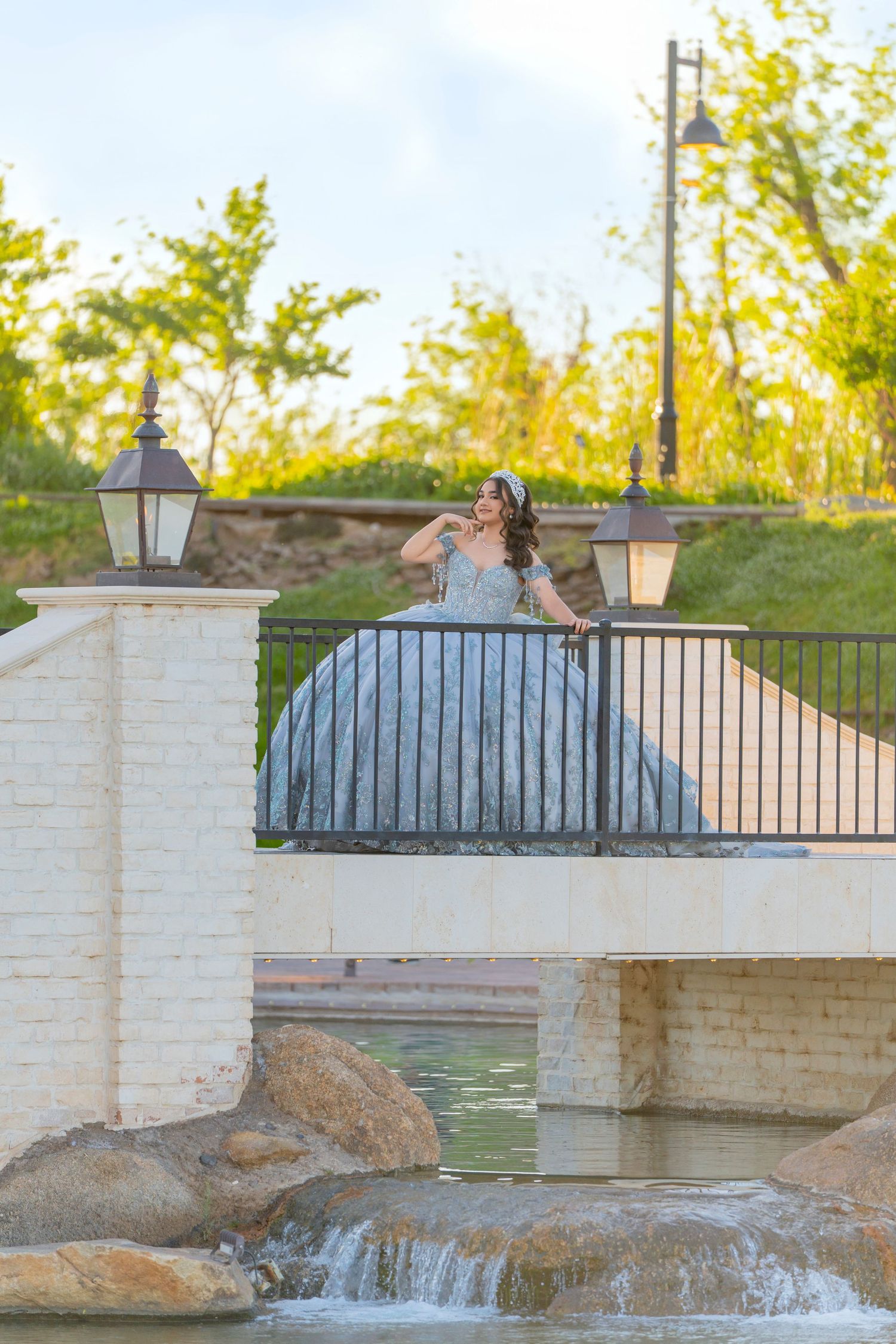 Young woman in elegant silver ball gown poses on decorative bridge with waterfall and lush greenery backdrop.