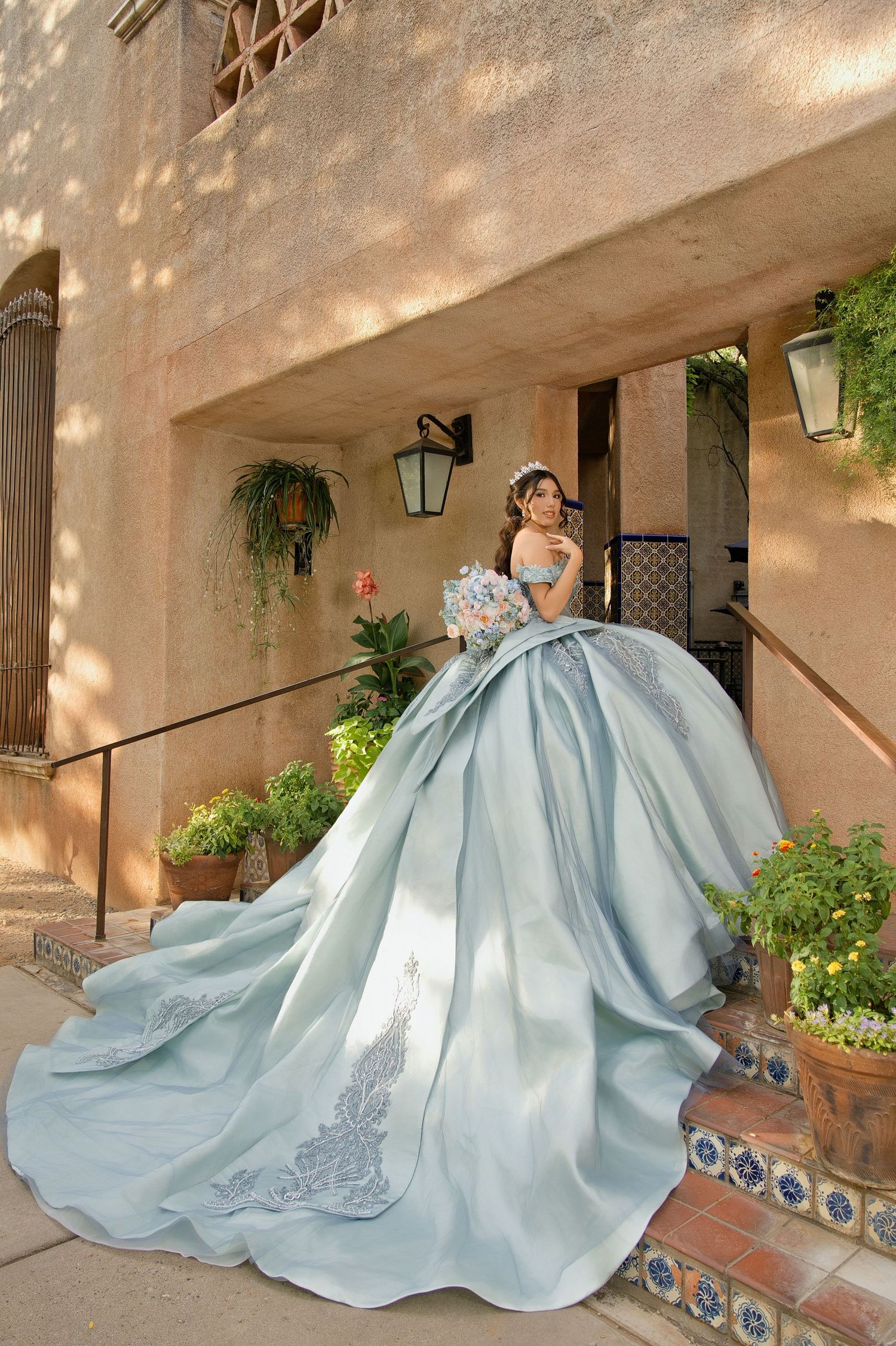 Woman in a stunning light blue ball gown with lace details poses on stone steps at an elegant outdoor venue.
