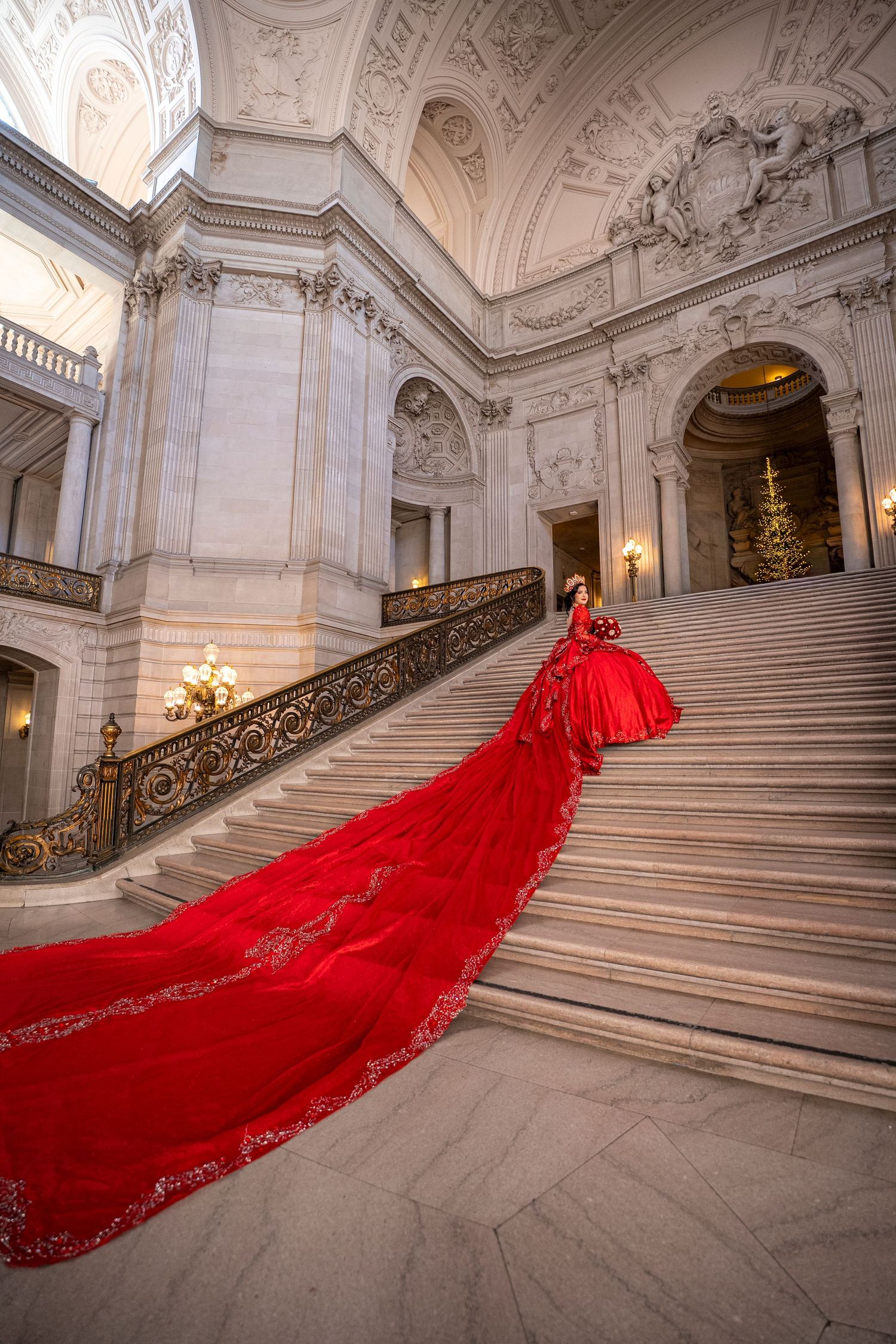 Woman in stunning red ballgown with long train descends grand marble staircase in ornate classical building.