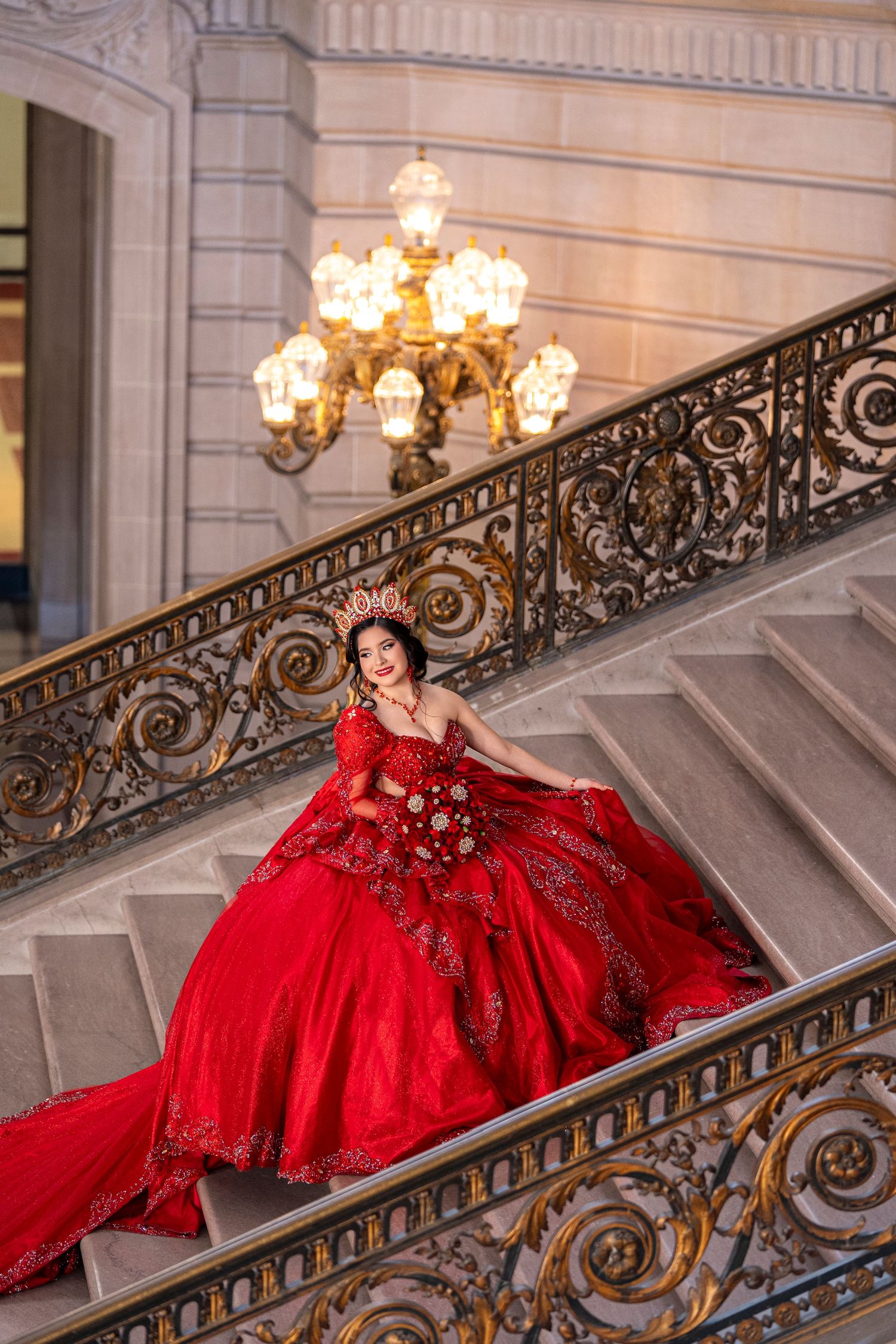Young woman in a stunning red quinceañera ball gown poses gracefully on ornate staircase under a golden chandelier.