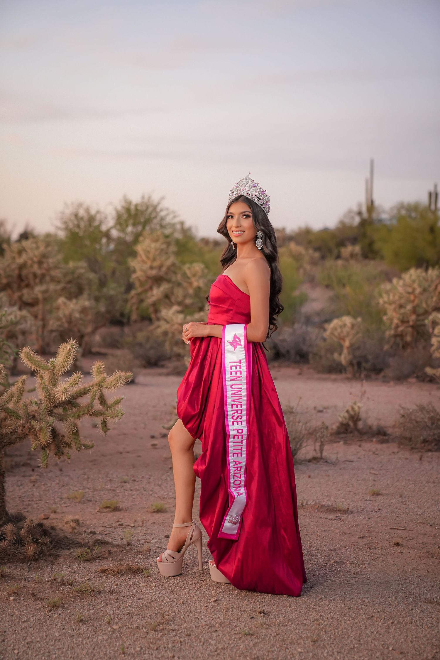 Woman in red gown and crown wearing a Miss Universe pageant sash poses in desert landscape at sunset.