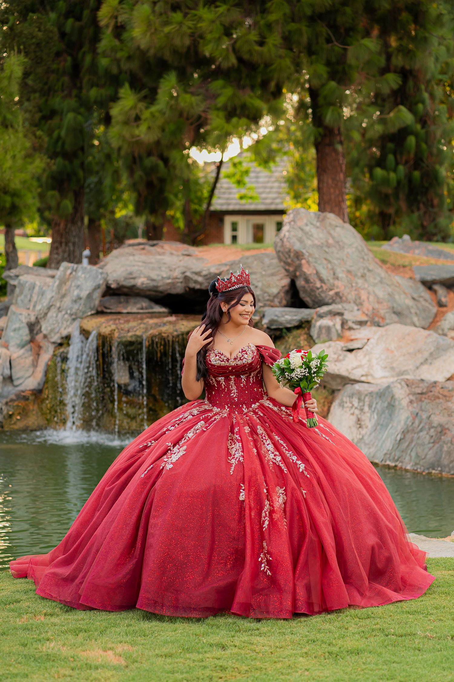 Young woman in red quinceañera ball gown with crown and bouquet poses by waterfall in lush garden.